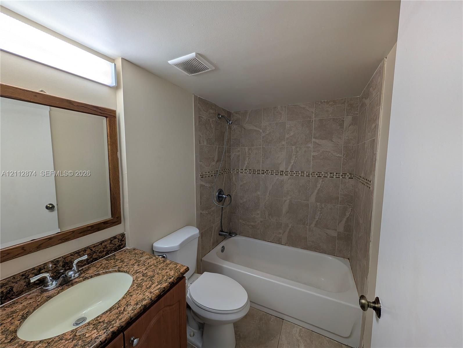 Vanity with speckled stone-finish countertop, integrated oval basin, framed wall mirror, and tub-shower combination featuring textured wall tile