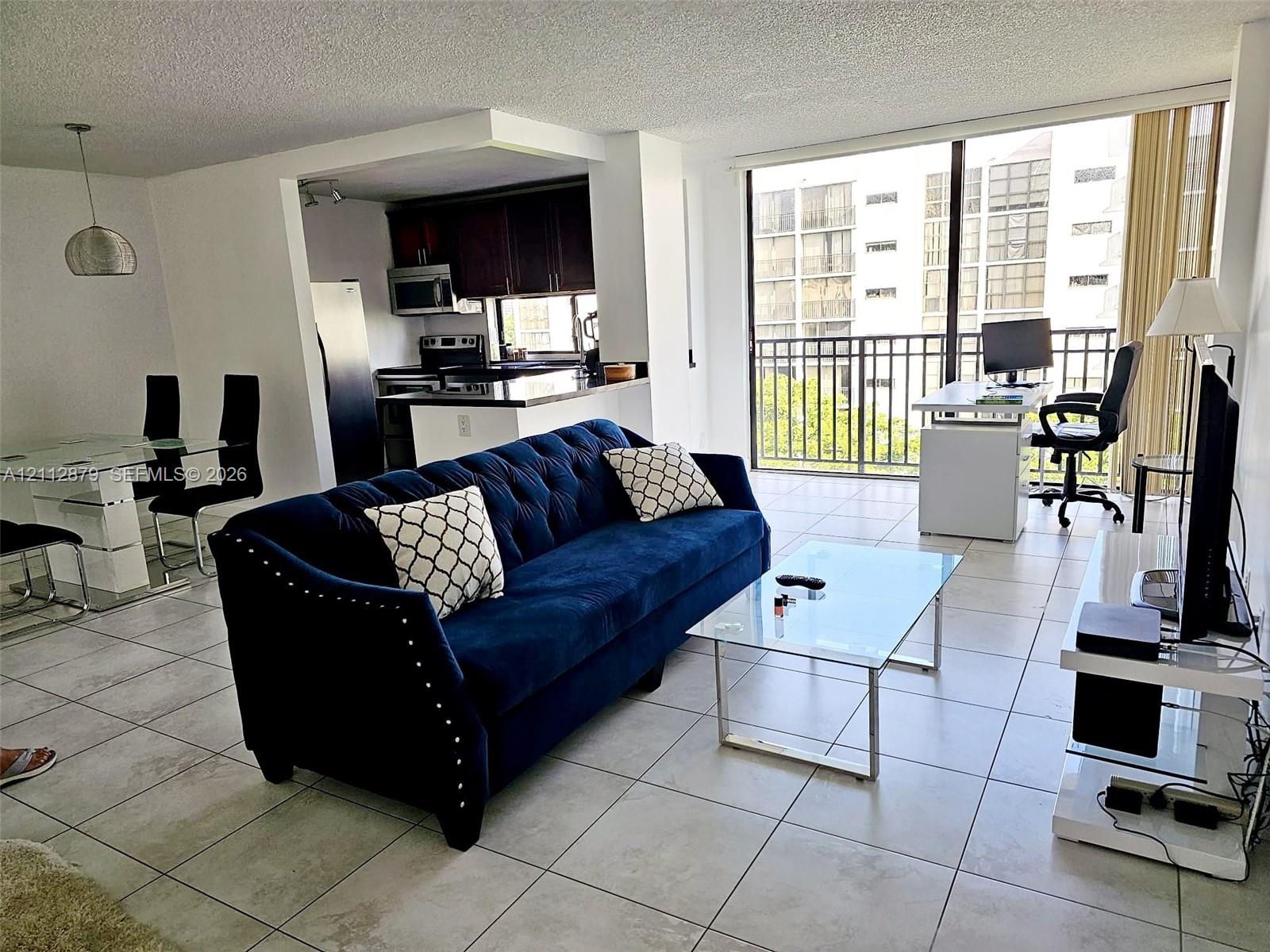 Open-concept floor plan with light-toned ceramic tile flooring, kitchen featuring dark wood cabinetry and a peninsula breakfast bar, floor-to-ceiling glass sliding doors, and a private metal-railing balcony