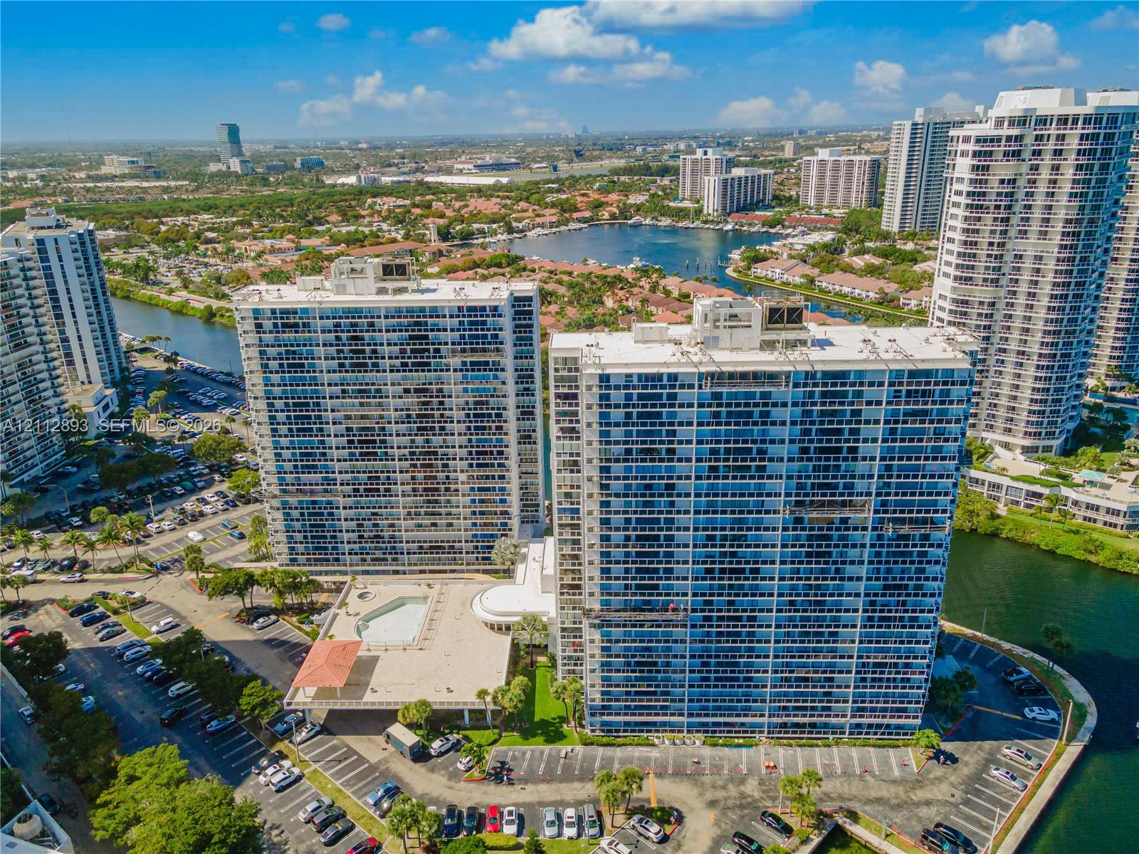 High-rise residential towers featuring expansive glass facades, a ground-level swimming pool, surface parking, and waterfront positioning