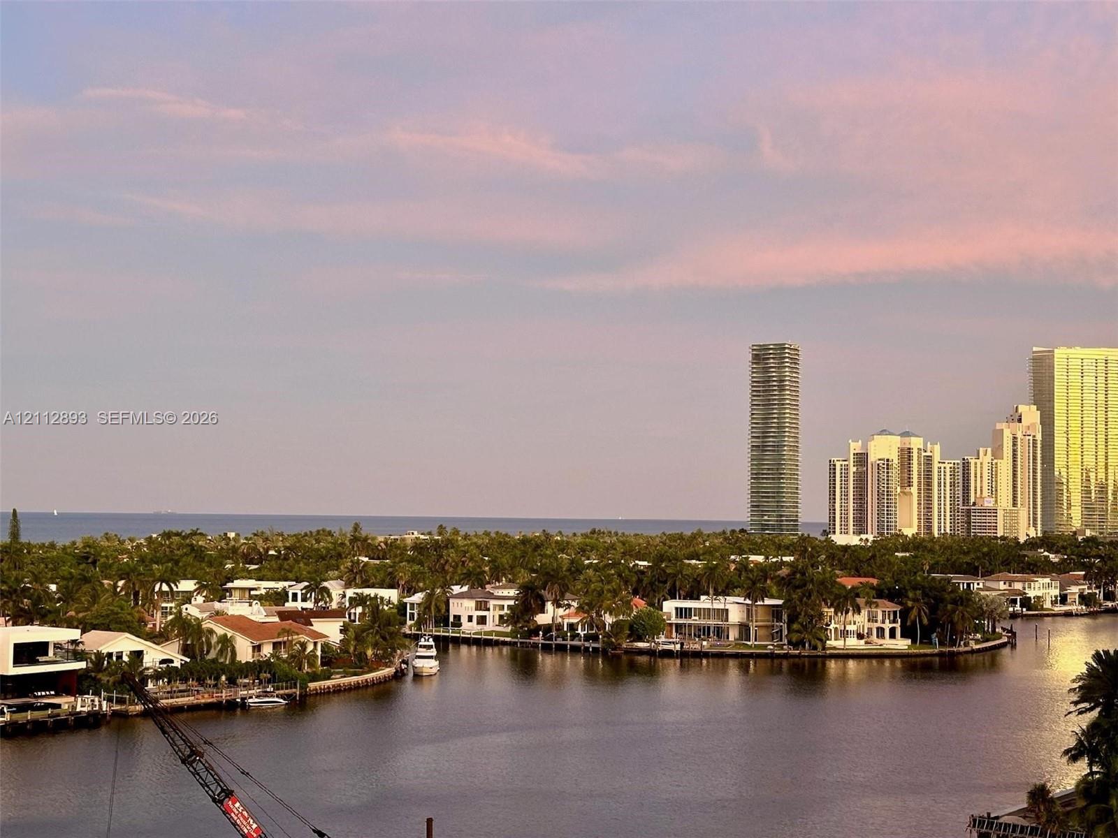 Waterfront landscape featuring a winding canal, mature palms, residential structures, and a distant coastal horizon