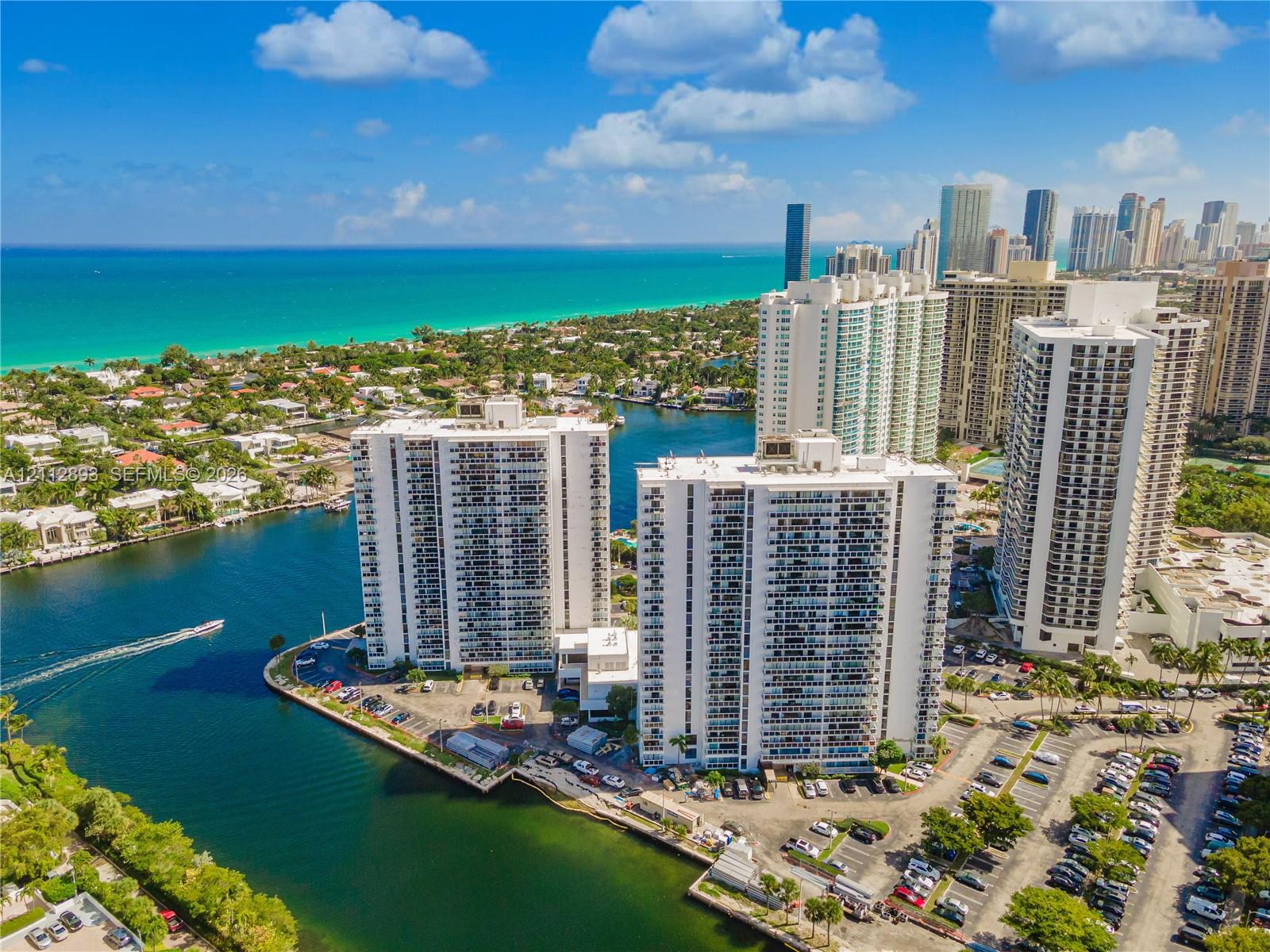 Aerial perspective of high-rise structures situated on a waterfront peninsula with direct coastal access and proximity to urban skyline, featuring on-site surface parking lots and established tropical landscaping