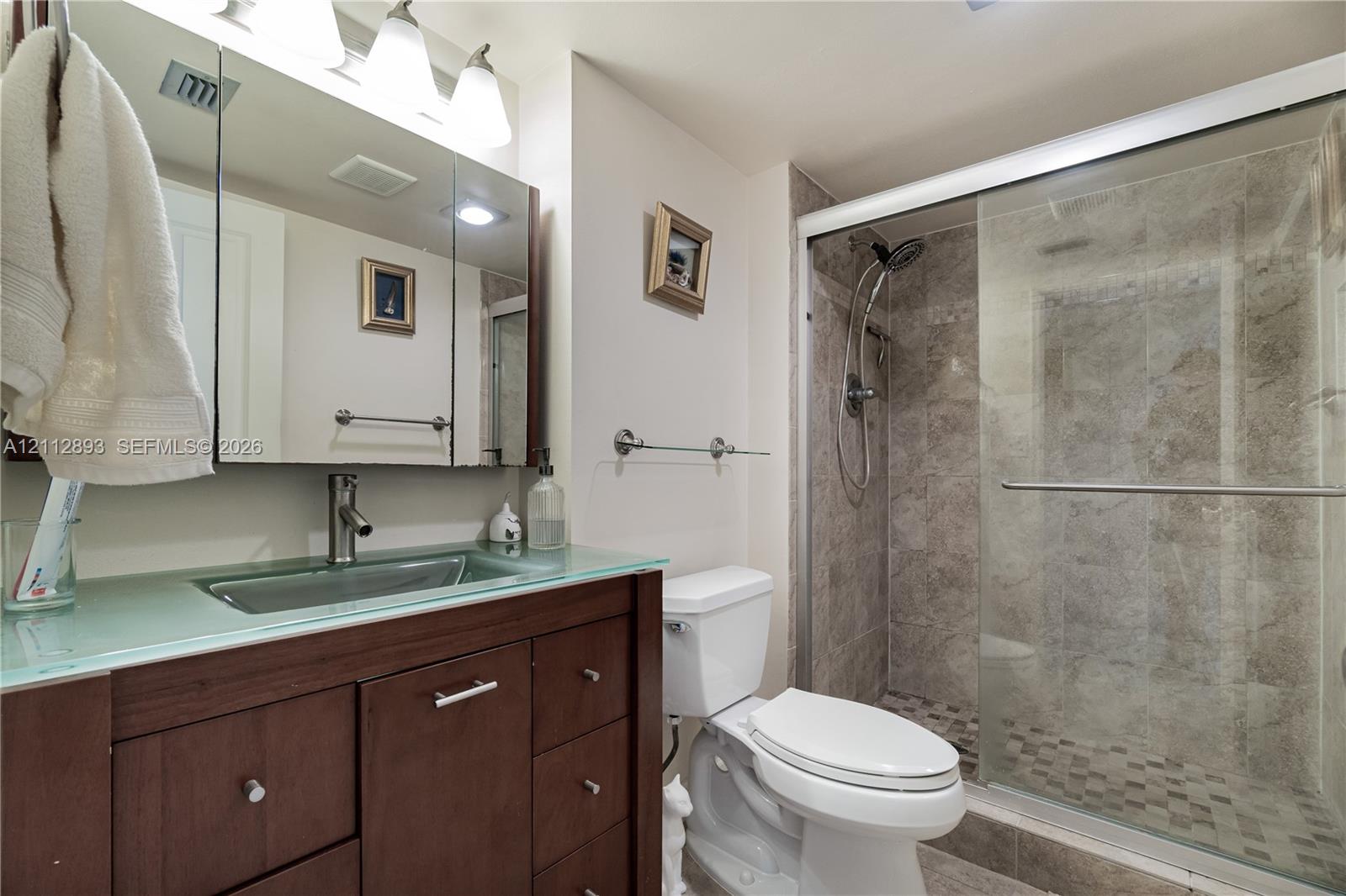 Vanity featuring a glass countertop with an integrated rectangular sink, dark wood-finish cabinetry, mirrored medicine cabinet, and a walk-in shower with tiled walls and glass sliding doors