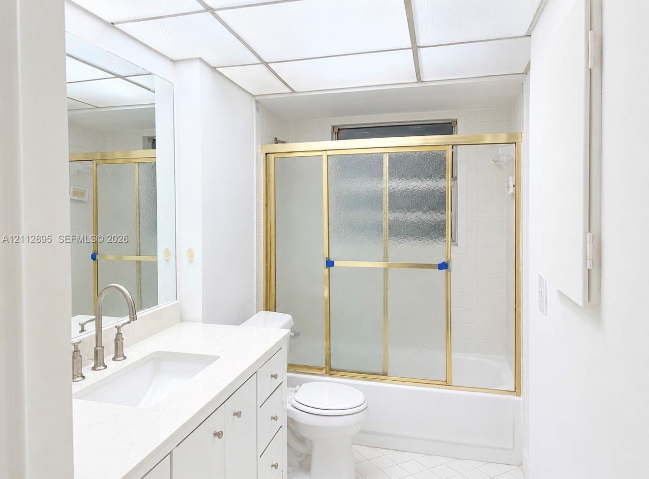Bathroom featuring a white vanity with a solid-surface countertop, an undermount sink, a drop-in tub with sliding glass doors, and a grid-style drop ceiling