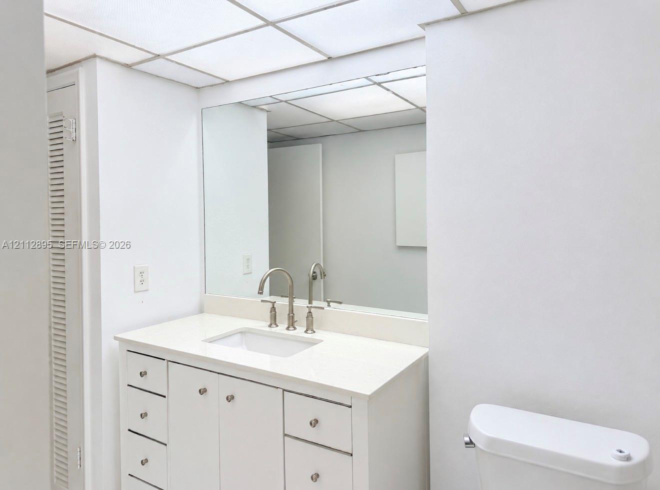 White vanity cabinet with integrated countertop, undermount rectangular sink, bridge-style faucet, large wall-mounted mirror, and drop ceiling grid lighting