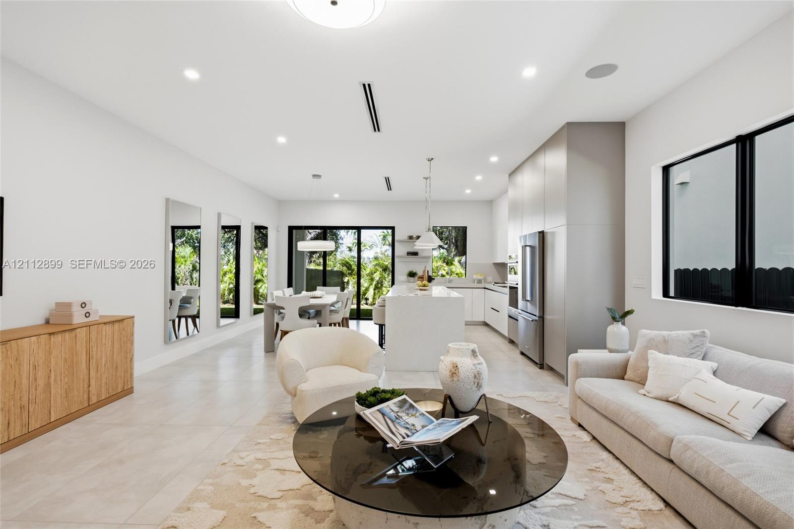 Open-concept interior featuring large-format tile flooring, recessed ceiling lighting, custom flat-panel cabinetry, a central kitchen island with waterfall-edge countertop, and sliding glass doors