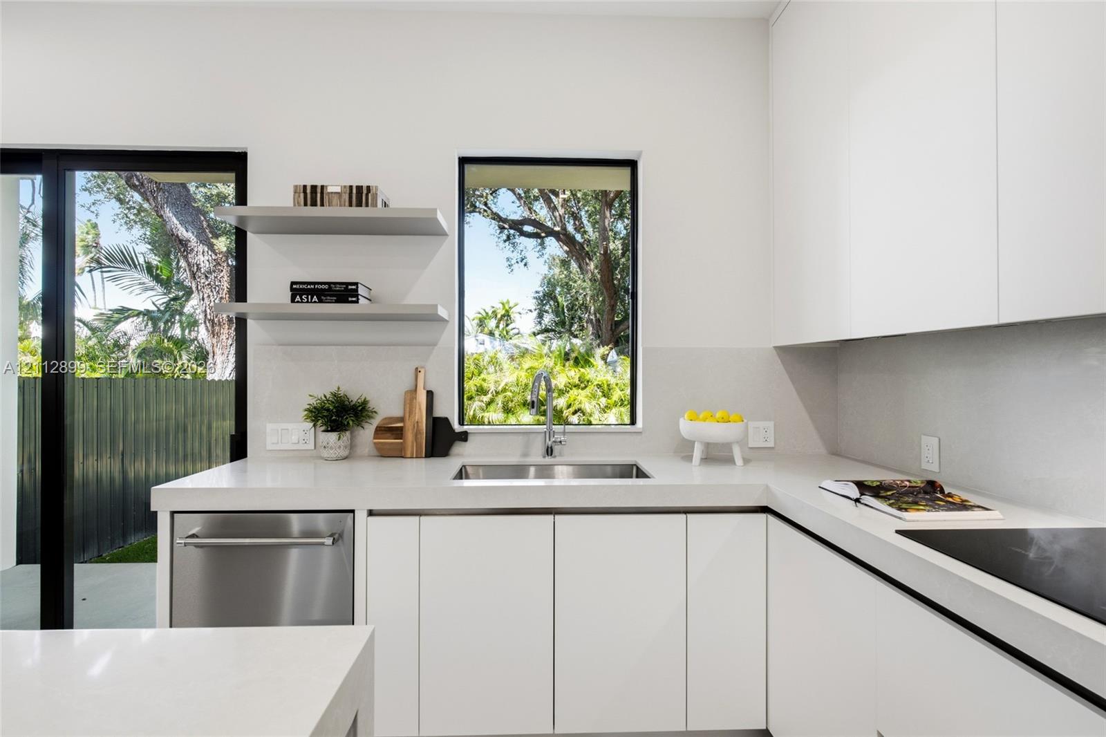 Minimalist kitchen featuring matte white flat-panel cabinetry, light-toned stone-surface countertops, stainless steel undermount sink, built-in dishwasher, and floating wall shelves