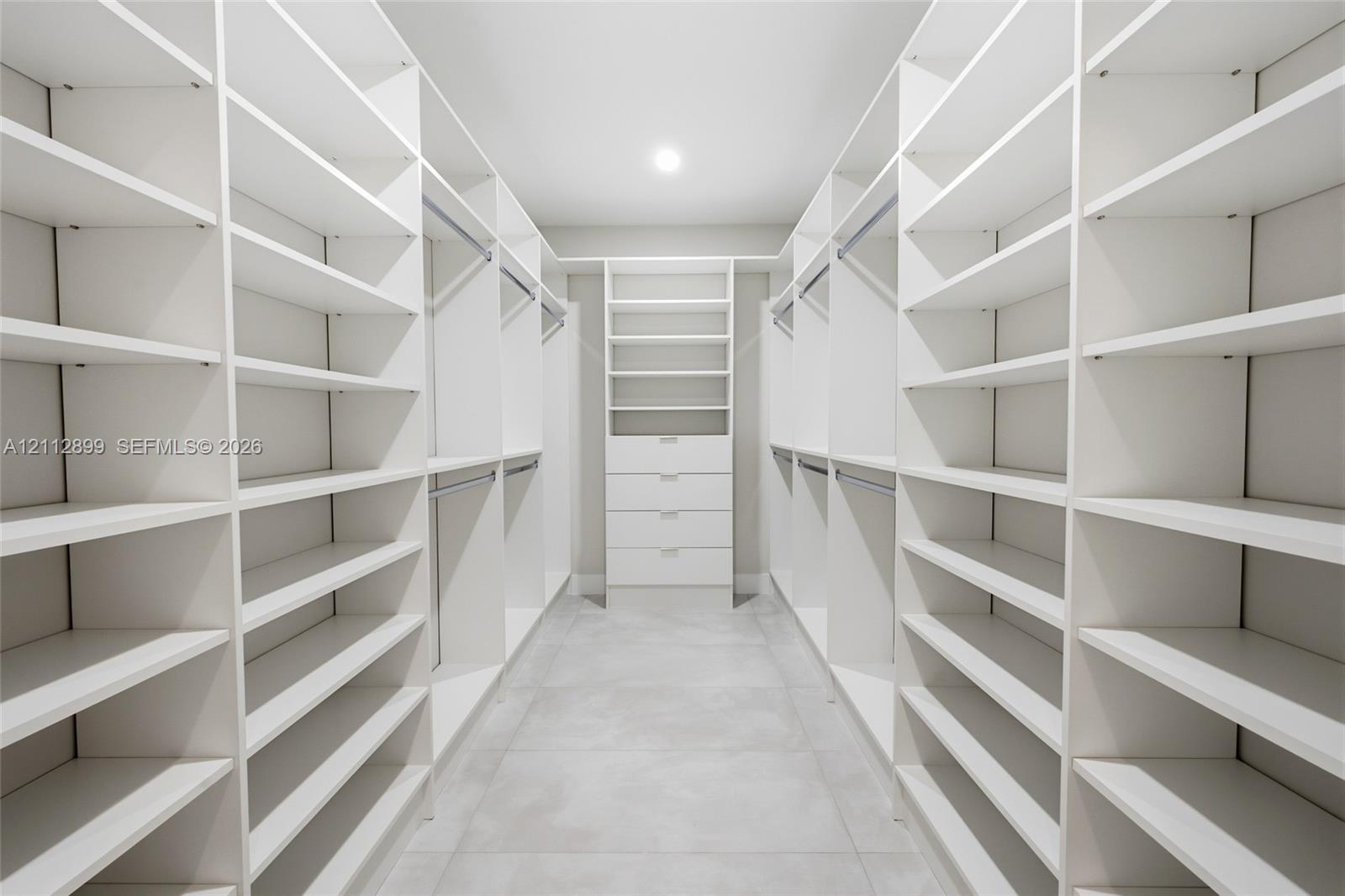 Walk-in closet featuring extensive built-in white shelving, integrated clothing rods, a central drawer bank, and tile flooring