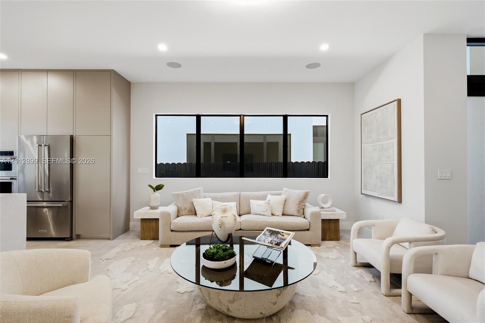 Open-concept living space featuring floor-to-ceiling cabinetry, stainless steel refrigerator, recessed lighting, and expansive tripartite black-framed window Open-concept living space featuring floor-to-ceiling cabinetry, stainless steel refrigerator, recessed lighting, and expansive tripartite black-framed window