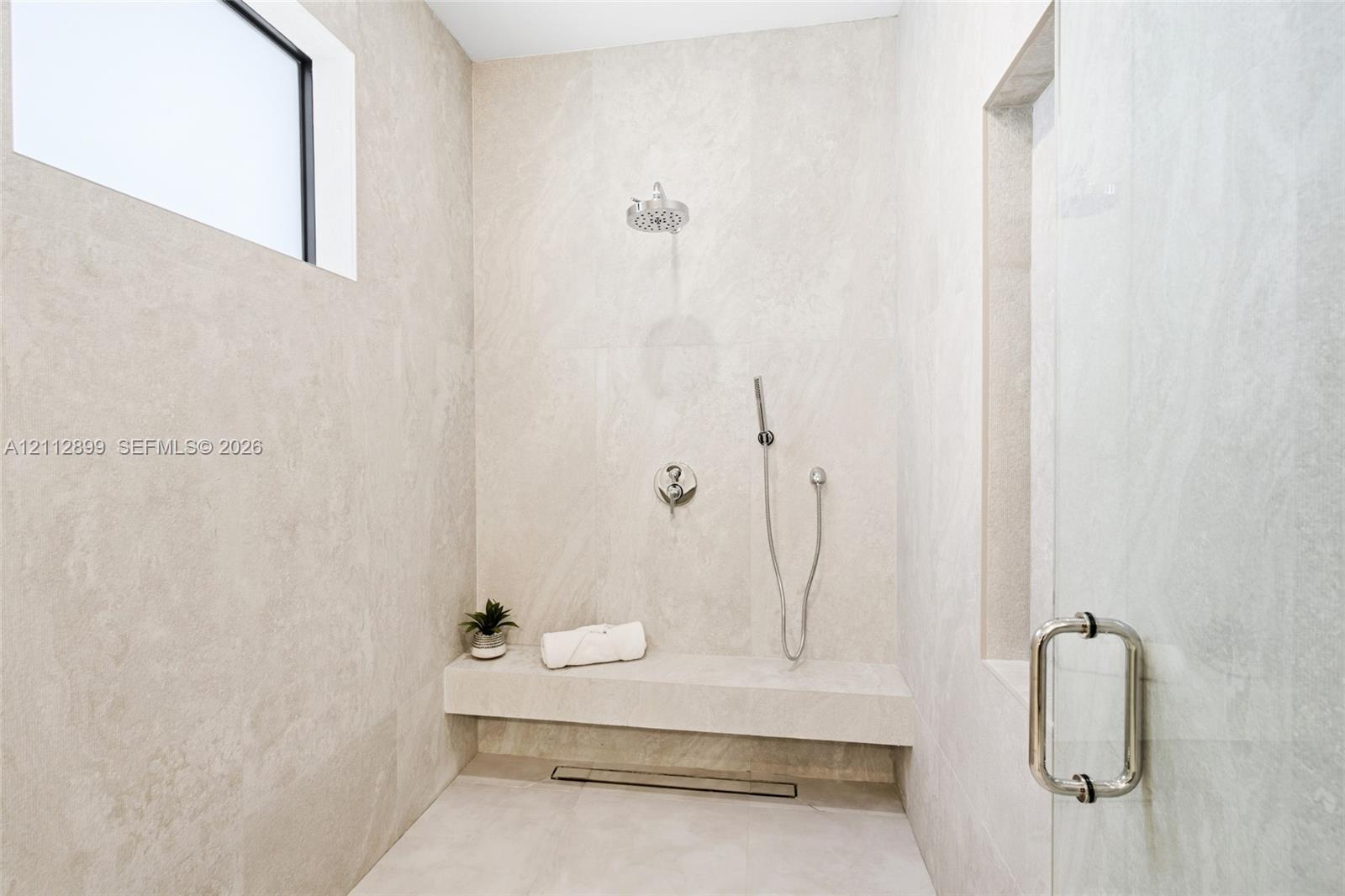 Walk-in shower featuring floor-to-ceiling stone-finish tile, a built-in stone bench, overhead rainfall showerhead, adjustable handheld shower wand, and glass enclosure door
