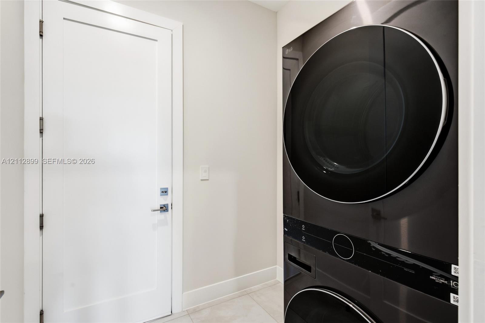 Stacked washer and dryer unit with a sleek black finish, paired with a white Shaker-style door featuring polished chrome hardware and large-format tile flooring