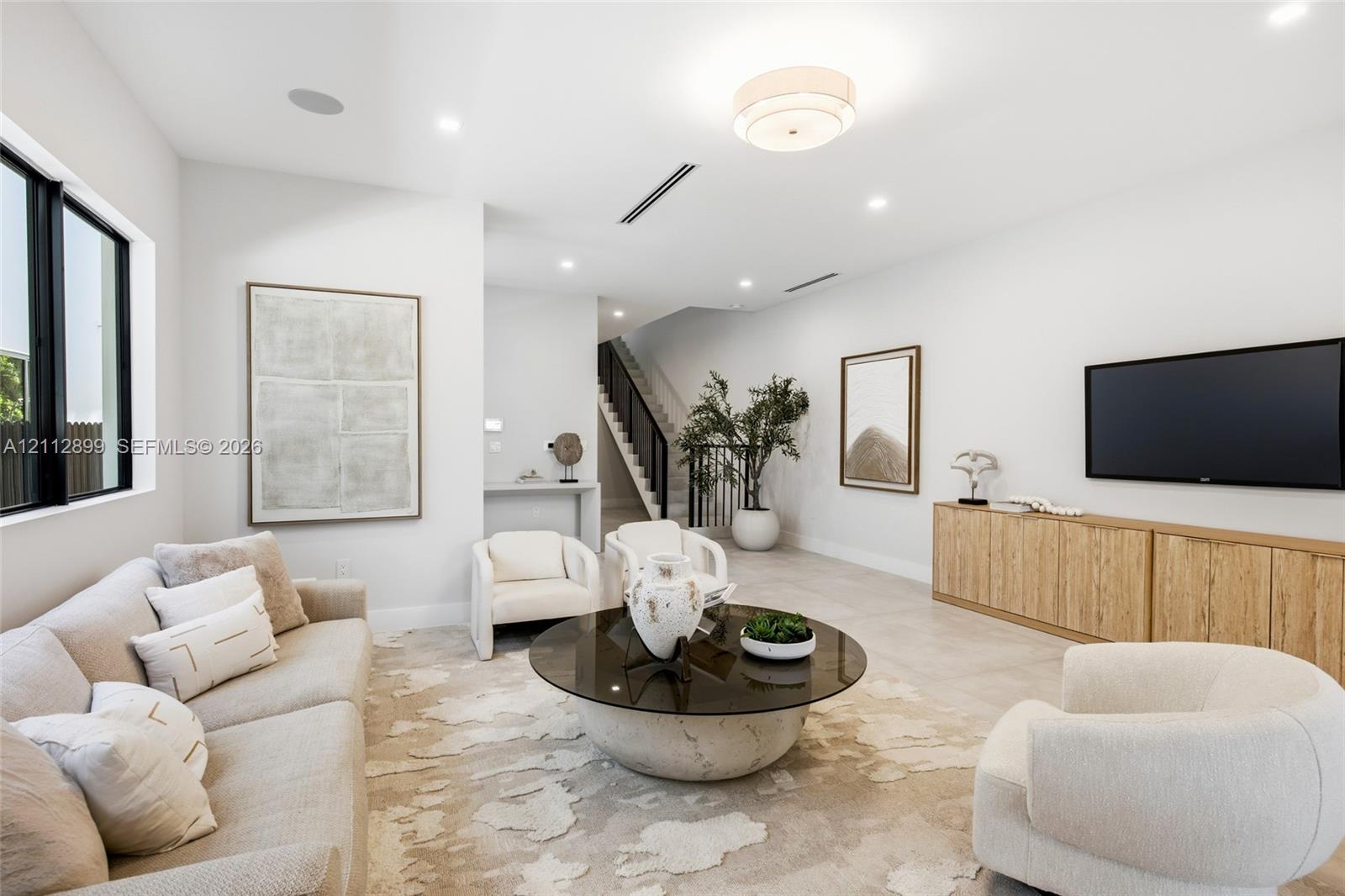 Open-concept living space featuring neutral oversized tile flooring, recessed ceiling lighting, a contemporary flush-mount fixture, black-framed windows, and built-in wood-finish cabinetry