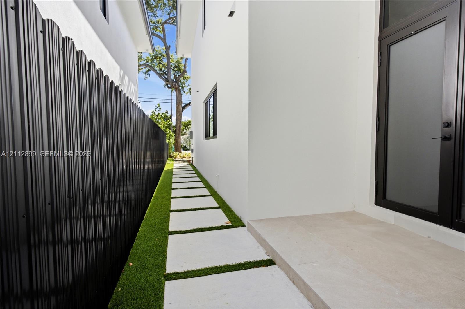 Side exterior featuring white stucco siding, a concrete paver walkway set in artificial turf, a black metal corrugated privacy fence, and a glass-panel entry door