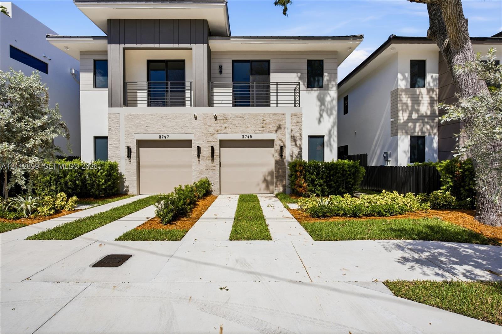 Modern exterior featuring a stacked stone facade, metal-accented second-story balcony with railings, dual two-car garage doors, and concrete paver driveways with integrated grass strips Modern exterior featuring a stacked stone facade, metal-accented second-story balcony with railings, dual two-car garage doors, and concrete paver driveways with integrated grass strips