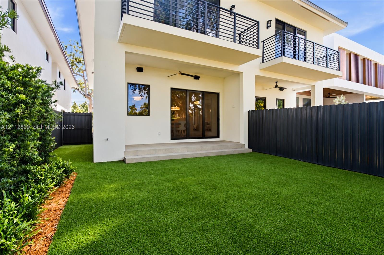 Rear exterior featuring a concrete patio, synthetic turf lawn, privacy fencing, and second-level balcony with metal railings