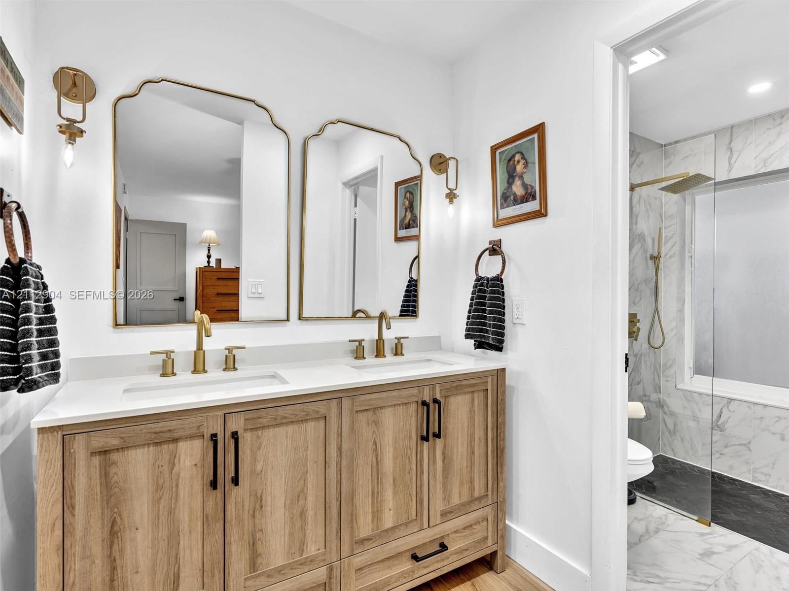 Dual-sink vanity with wood-finish cabinetry, quartz countertop, brass fixtures, arched metal-frame mirrors, and a glass-enclosed shower