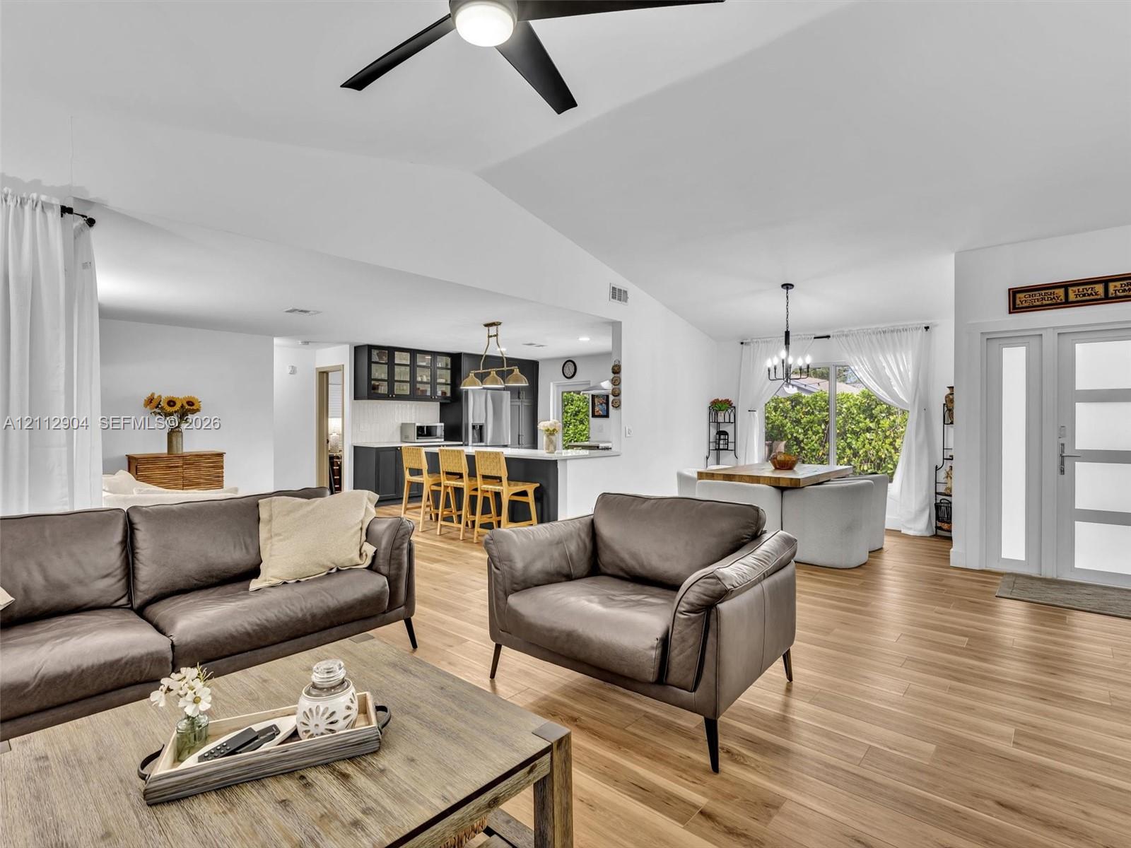 Open-concept interior featuring vaulted ceilings, wood-finish flooring, a kitchen island with built-in cabinetry, a multi-pane glass entry door, and a modern ceiling fan Open-concept interior featuring vaulted ceilings, wood-finish flooring, a kitchen island with built-in cabinetry, a multi-pane glass entry door, and a modern ceiling fan