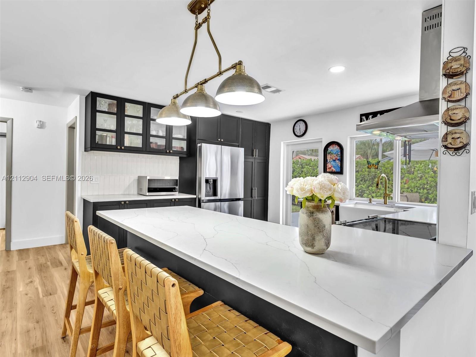 Kitchen featuring dark cabinetry with glass-panel upper doors, a large kitchen island with a stone-finish countertop, stainless steel appliances, a multi-pendant light fixture, and wood-finish flooring