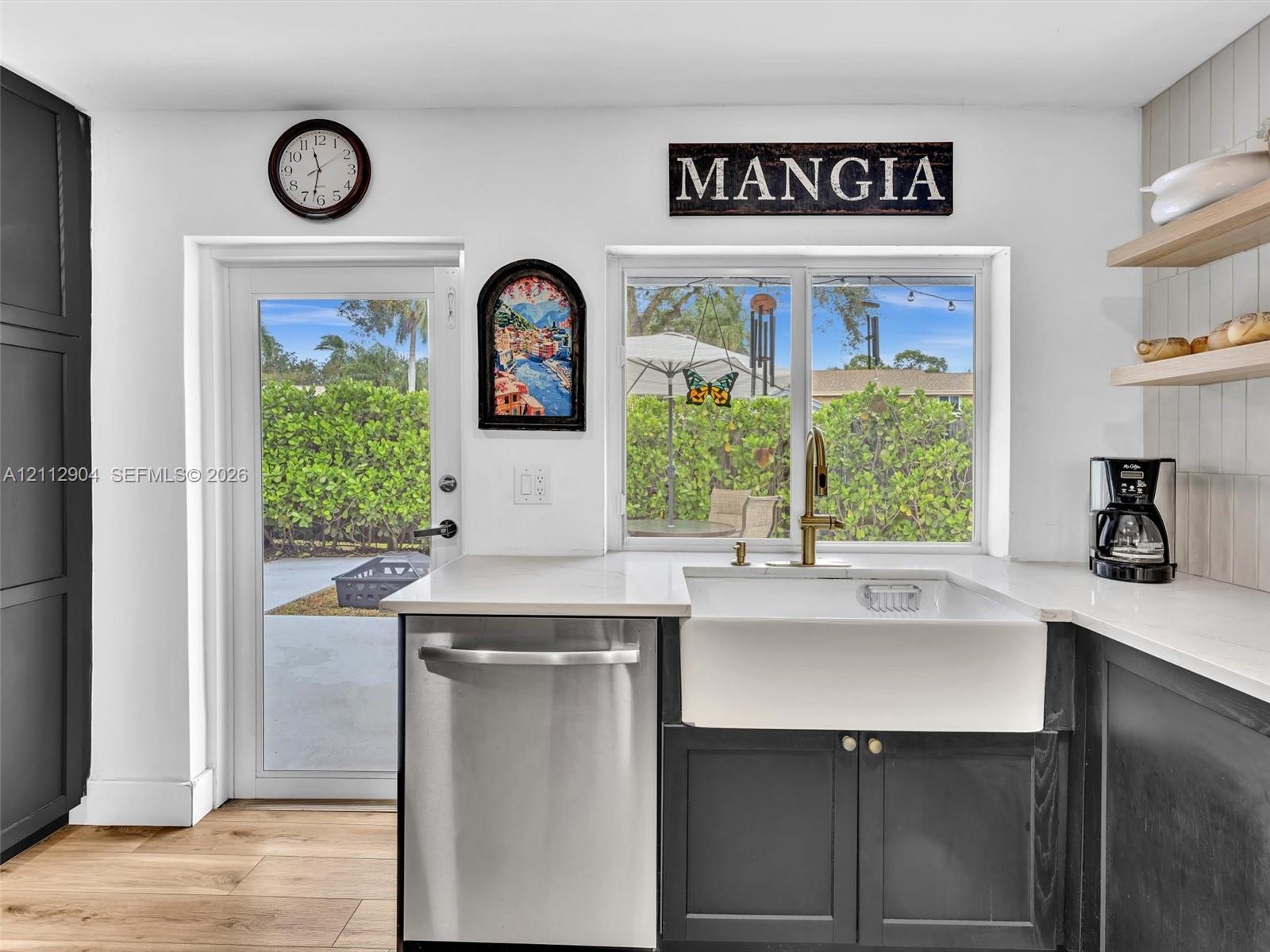 Kitchen featuring charcoal-toned lower cabinetry, white apron-front sink with gold-tone fixture, light-toned quartz countertops, wood-finish flooring, and floating wood shelving