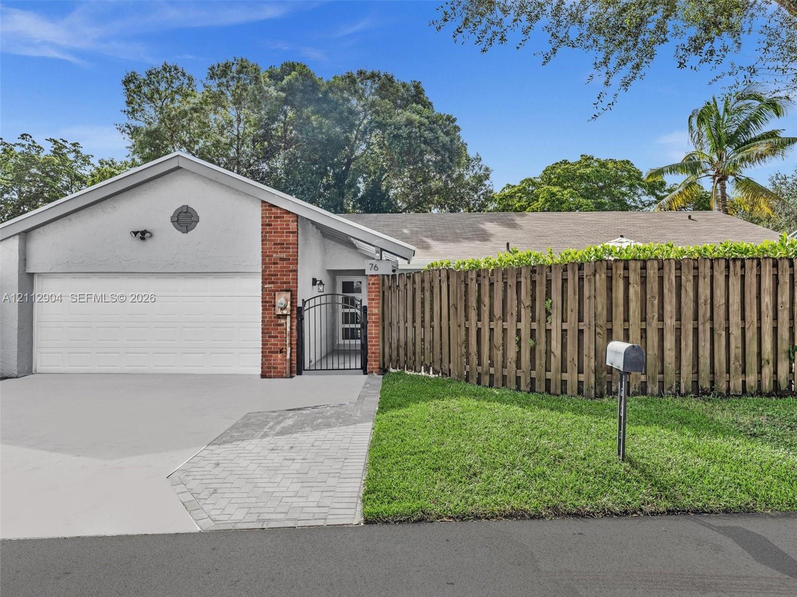 Exterior facade featuring a stucco finish, brick accents, attached garage, paved walkway, and wood-slat privacy fencing