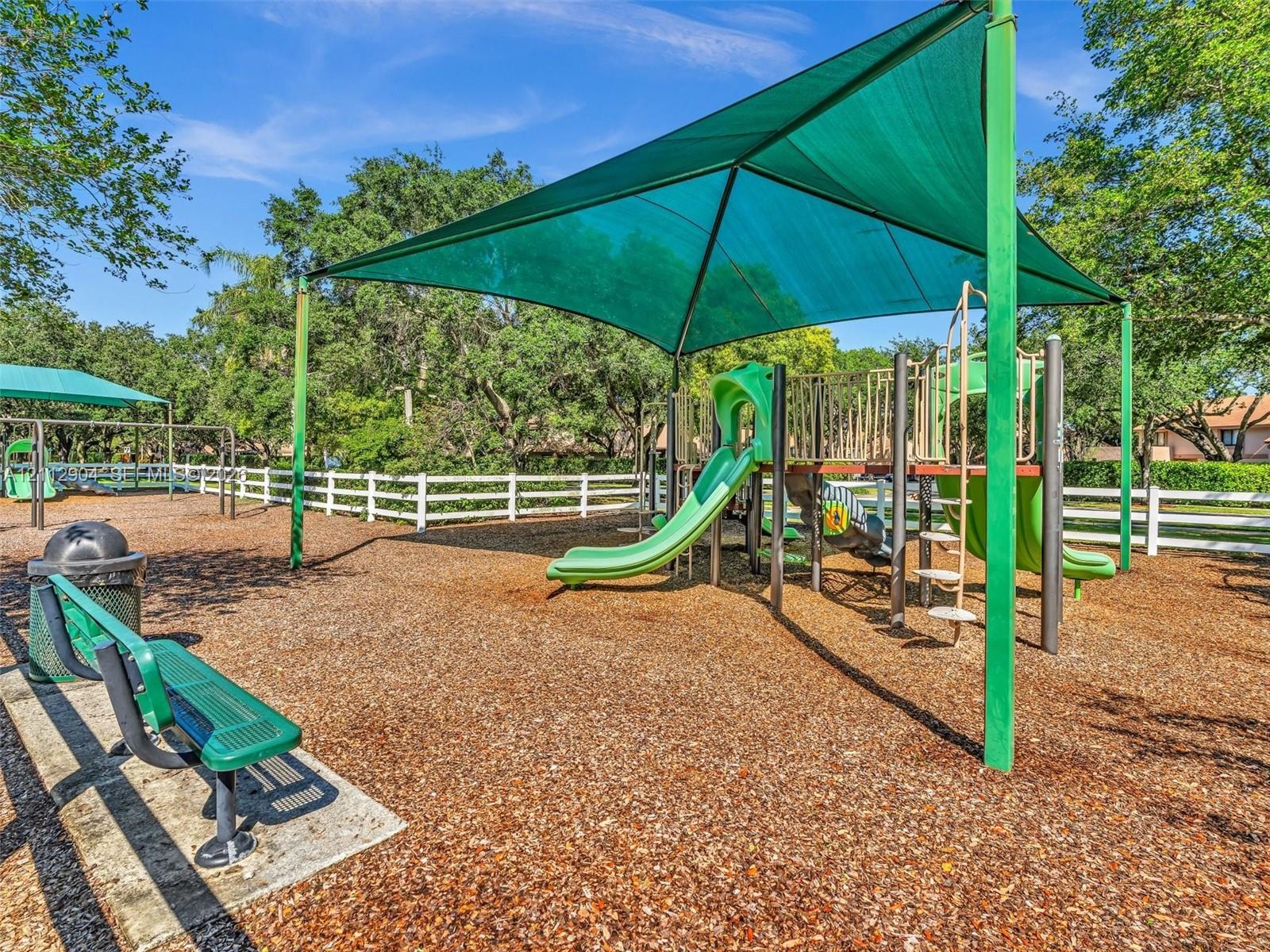 Recreational area featuring a modular playground structure, shade canopy, integrated metal bench, and white perimeter fencing
