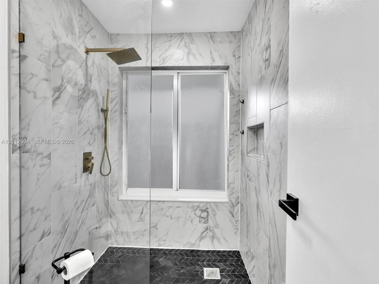 Walk-in shower featuring large-format stone-finish wall tile, herringbone-patterned floor tile, a recessed wall niche, brass-finish rainfall shower head with handheld attachment, and a glass partition