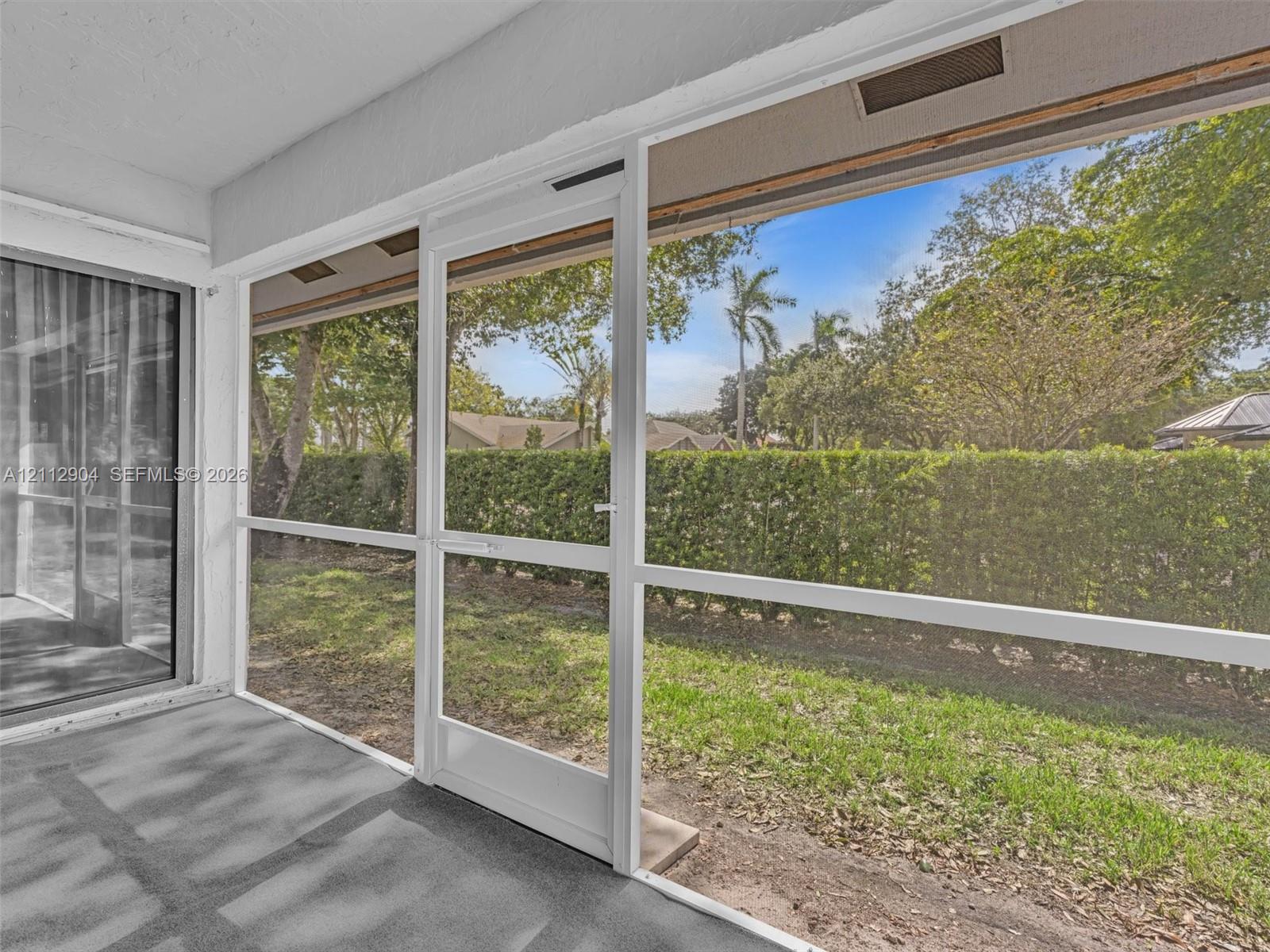 Screened-in porch featuring a concrete floor, white aluminum framing, and a screen door, opening to a grass yard with a perimeter privacy hedge