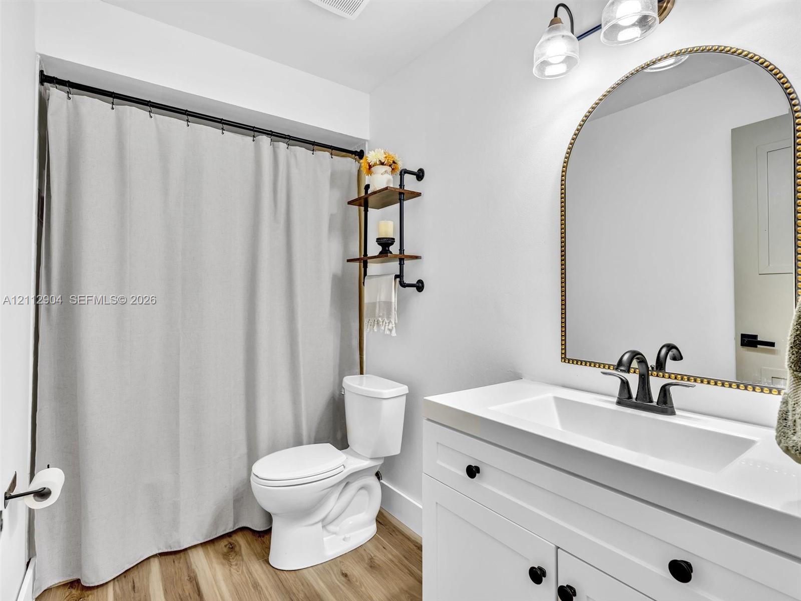 Bathroom featuring a white vanity with an integrated rectangular sink, matte black hardware, an arched mirror with metallic trim, wood-finish flooring, and industrial-style shelving