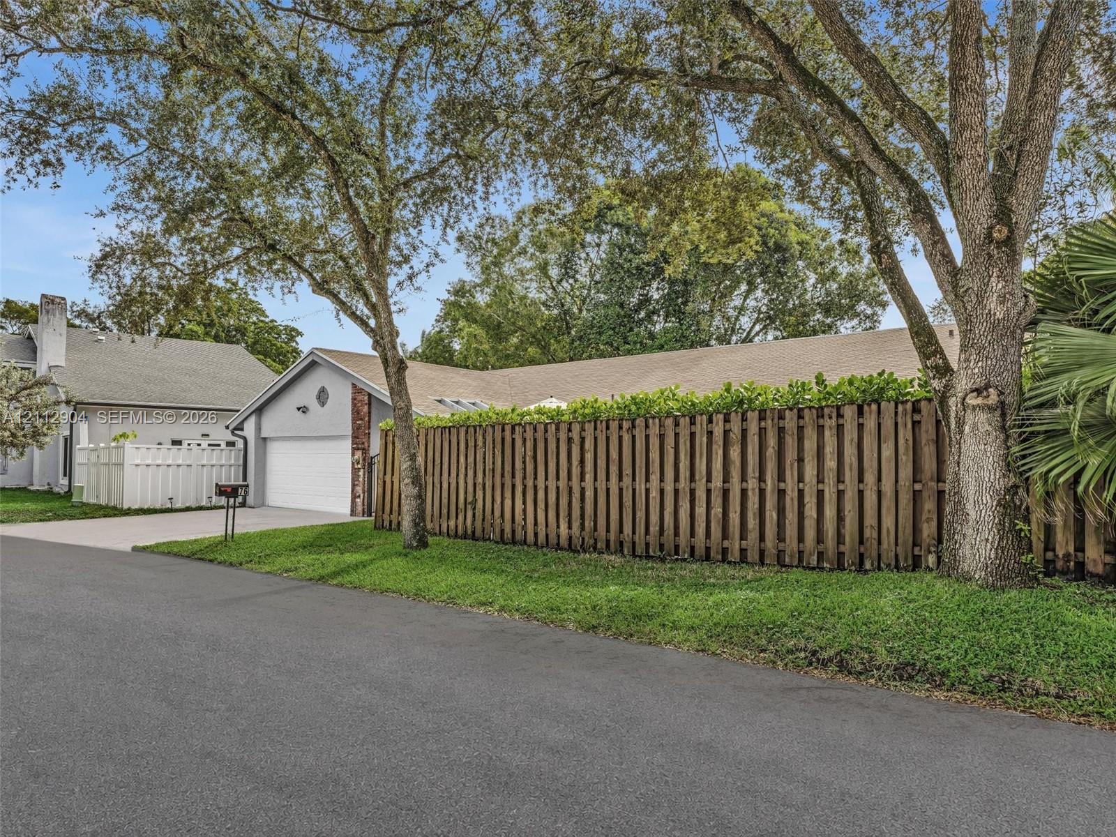 Exterior facade featuring a concrete driveway, two-car garage, shingle roofing, and vertical wood privacy fencing, complemented by mature trees and manicured landscaping