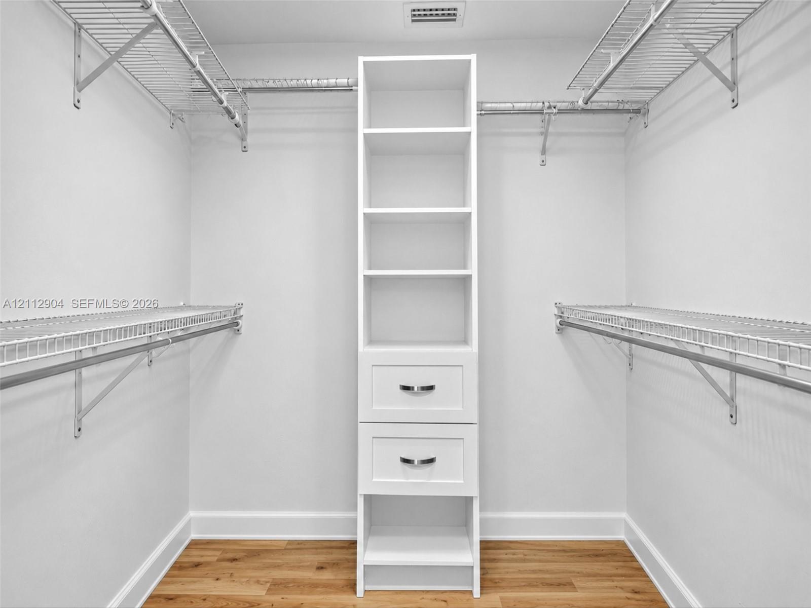 Walk-in closet featuring wood-finish flooring, white built-in cabinetry with two storage drawers, and dual-level metal wire shelving