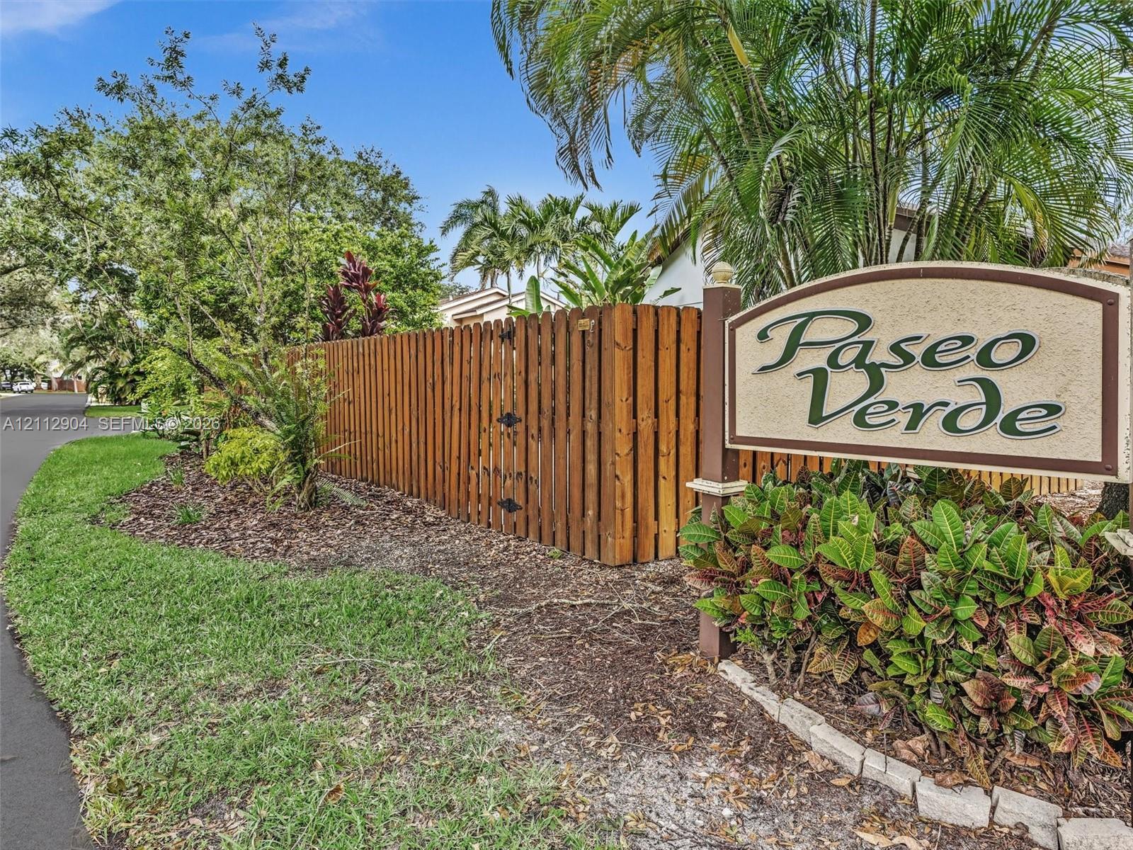 Paseo Verde community signage featuring a custom-carved monument, vertical wood-plank fencing, asphalt-paved thoroughfare, and manicured tropical landscaping