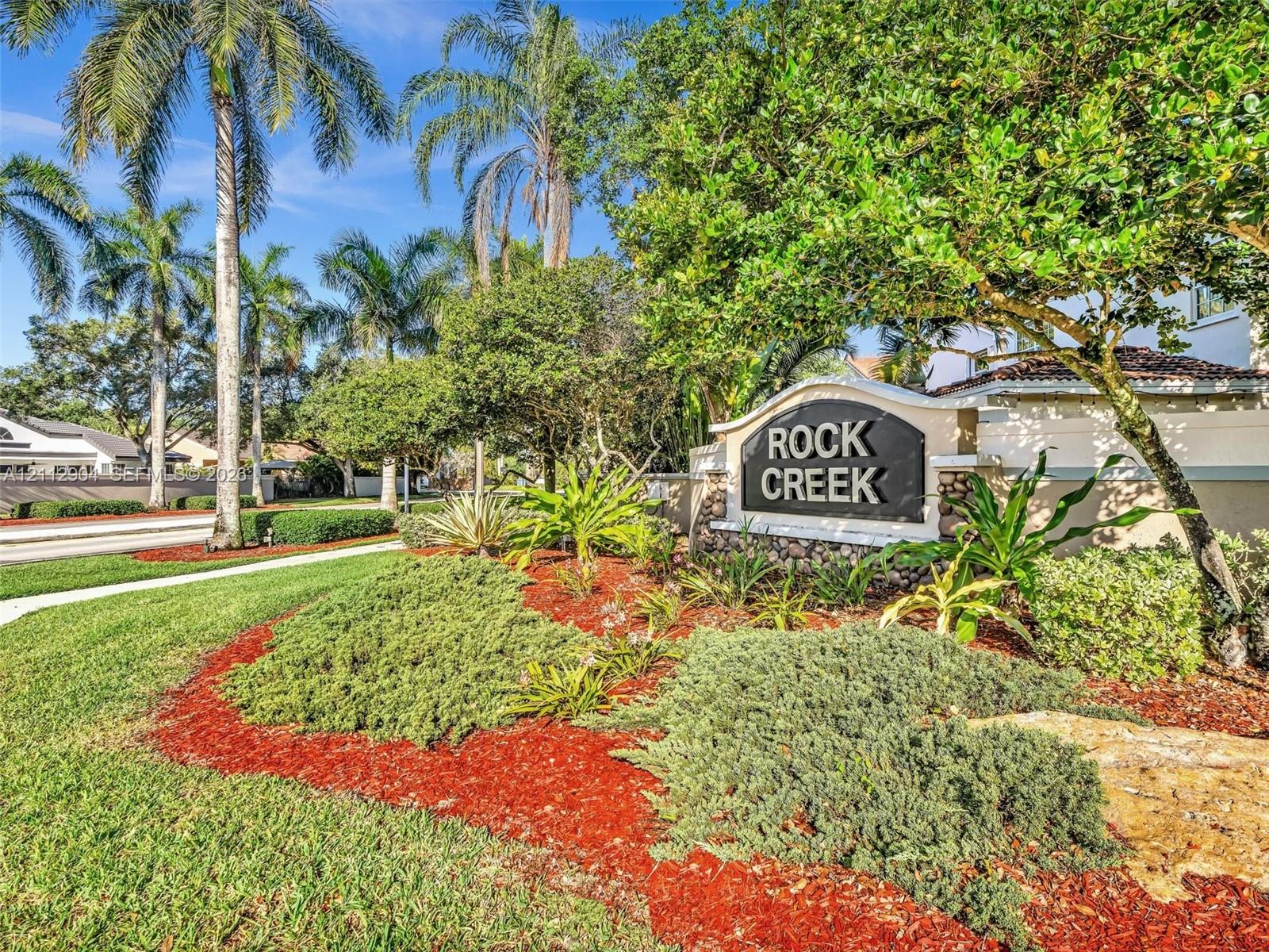 Community signage featuring stone-accented monument base, mature palm trees, established tropical landscaping, and expansive green space