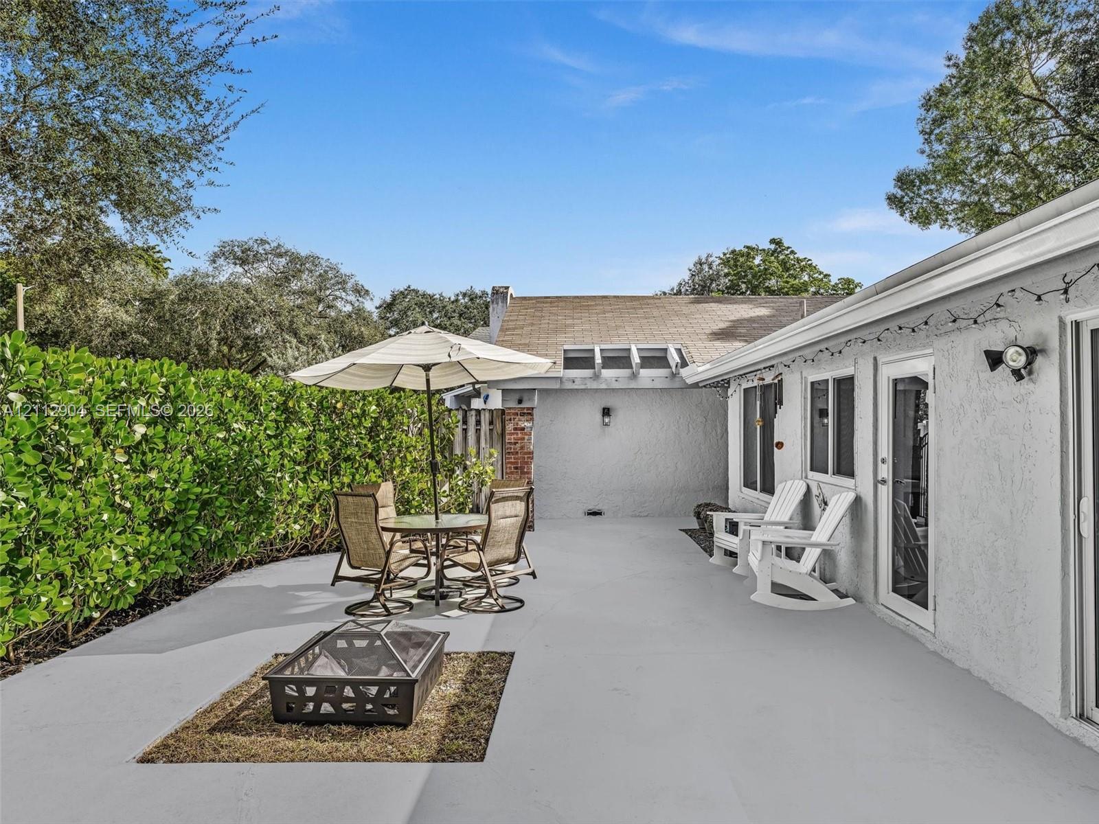 Expansive concrete patio featuring a dedicated fire pit area, stucco exterior, and tall privacy hedging