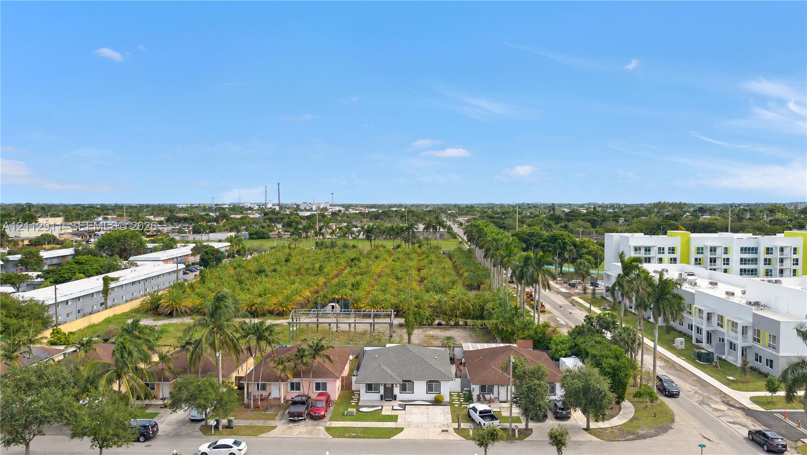 Aerial perspective featuring a single-story residence with a pitched roof, paved driveway, expansive level lot, and mature palm landscaping
