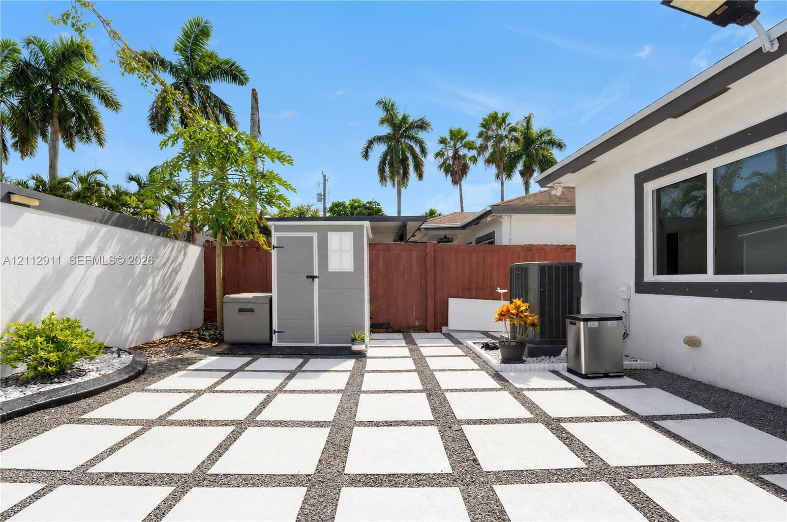 Rear outdoor area featuring a grid-patterned concrete paver patio with gravel fill, enclosed by a white stucco perimeter wall and wood privacy fencing