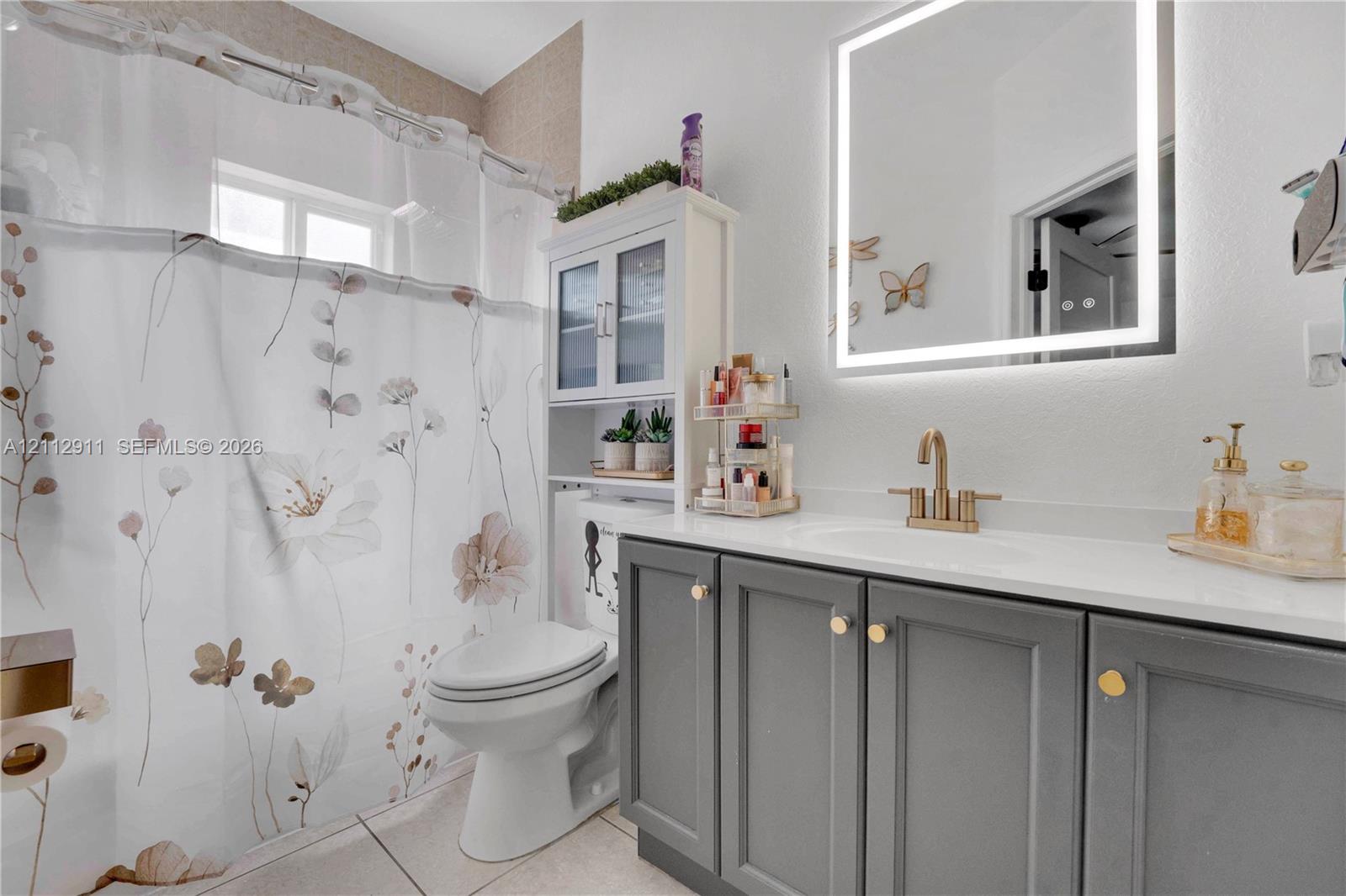 Bathroom featuring a gray shaker-style vanity with a solid-surface countertop, an integrated sink with a brass-finish faucet, and a wall-mounted LED-backlit mirror
