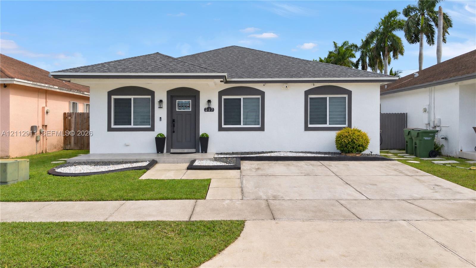 Single-story exterior featuring a white stucco facade, contrasting dark gray trim, a concrete driveway, and landscaped front beds with stone accents