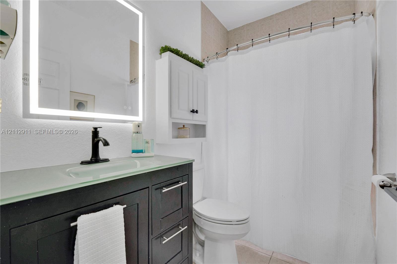 Dark-finish vanity with a glass countertop and integrated sink, matte black faucet, wall-mounted illuminated mirror, and white built-in storage cabinet