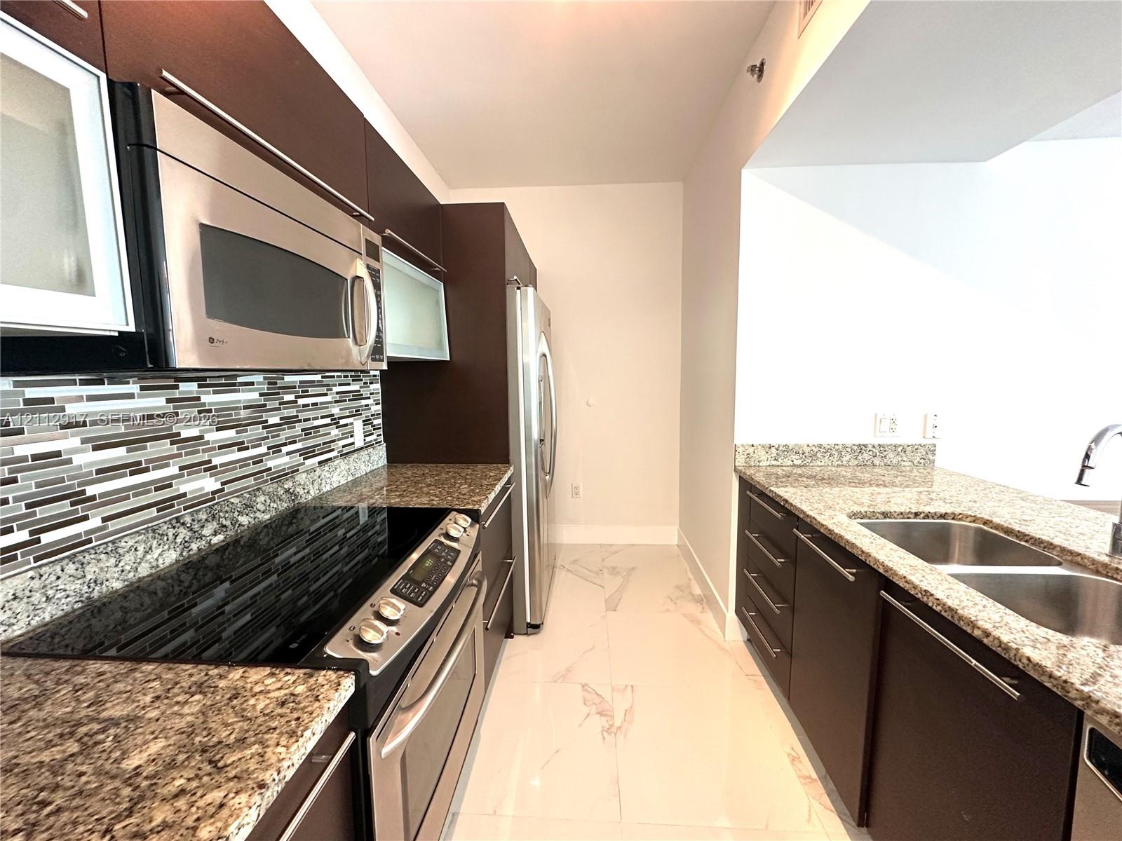 Kitchen featuring dark wood-finish cabinetry, stainless steel appliances, speckled stone countertops, mosaic tile backsplash, and light-toned tile flooring