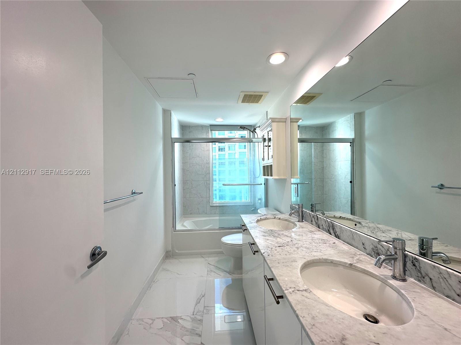 Bathroom featuring a dual-sink vanity with stone-surfaced countertops, expansive wall-to-wall mirror, recessed lighting, and a bathtub with a glass sliding enclosure