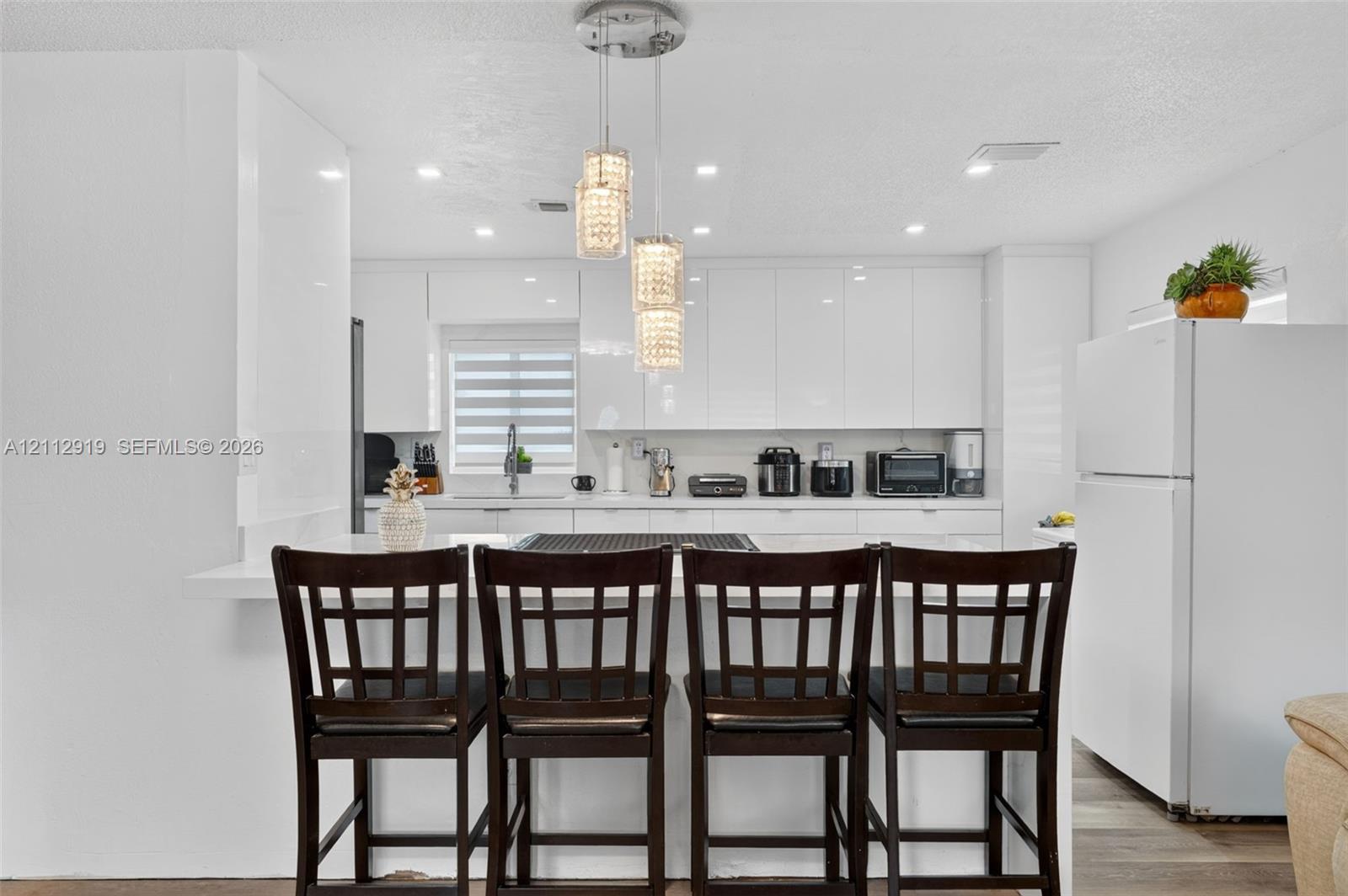 Kitchen featuring high-gloss white cabinetry, white countertops, a multi-tier crystal pendant light fixture, recessed lighting, and a breakfast bar
