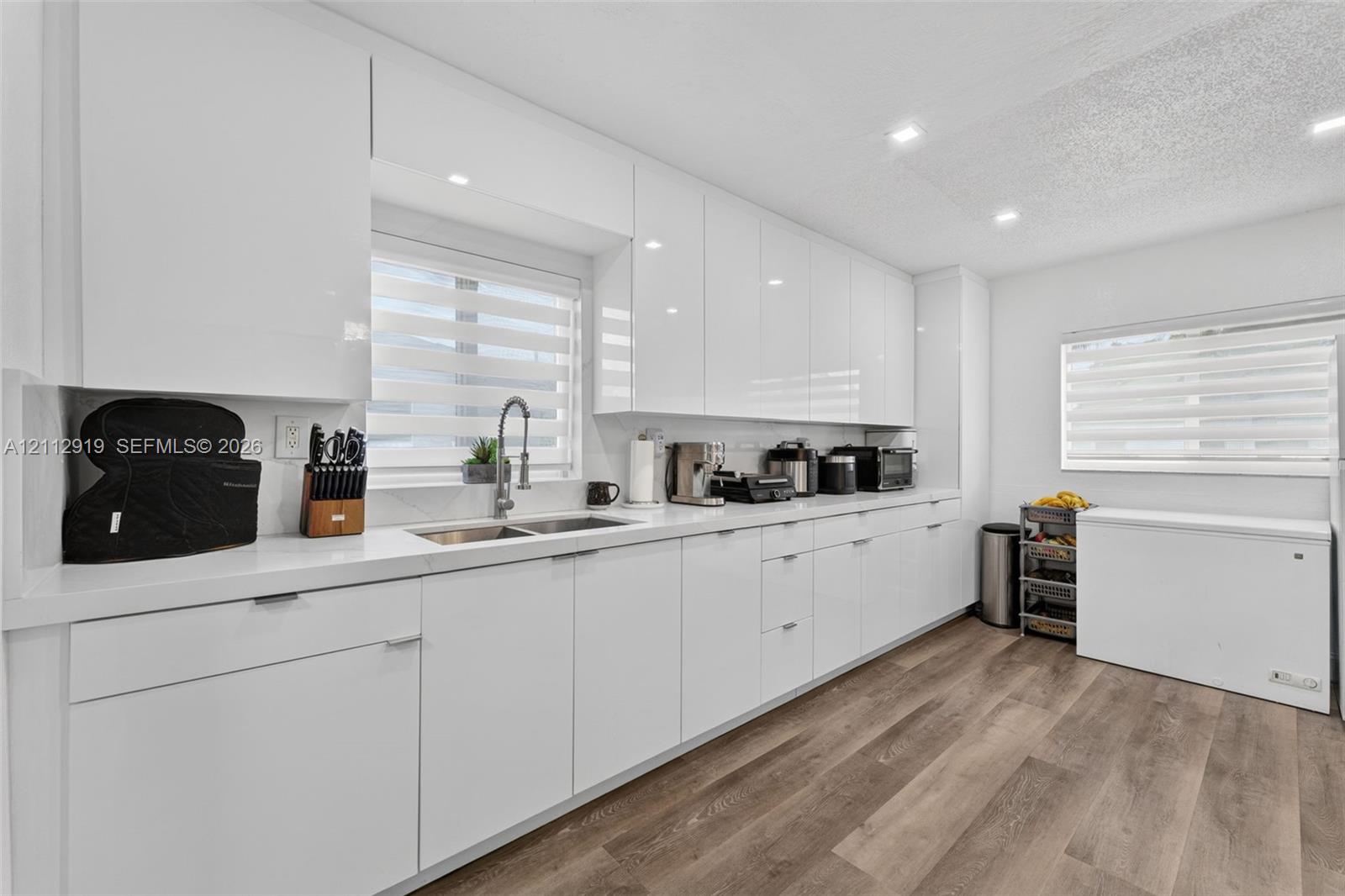 Modern kitchen featuring high-gloss white cabinetry, white stone-surface countertops, wood-finish flooring, undermount dual-basin stainless steel sink, and recessed ceiling lighting