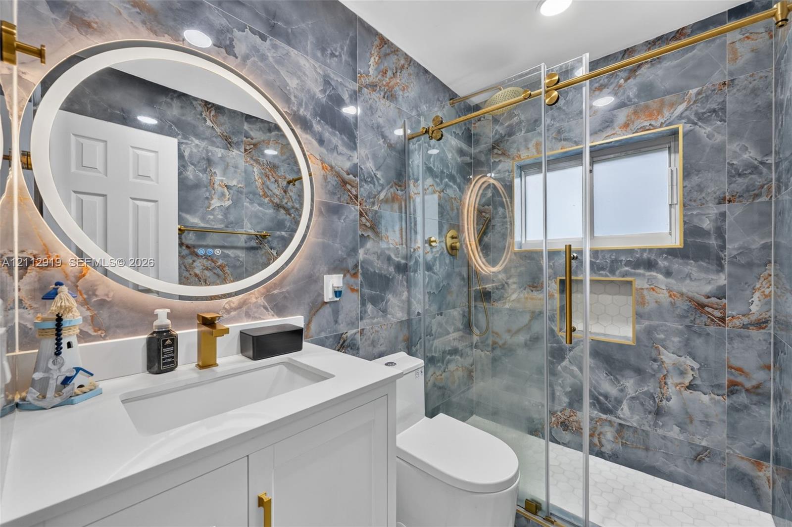 Bathroom featuring floor-to-ceiling blue veined stone-look wall tile, backlit circular vanity mirror, white vanity with rectangular under-mount sink, brass-finish fixtures, and a glass-enclosed shower with gold-tone hardware and built-in niche