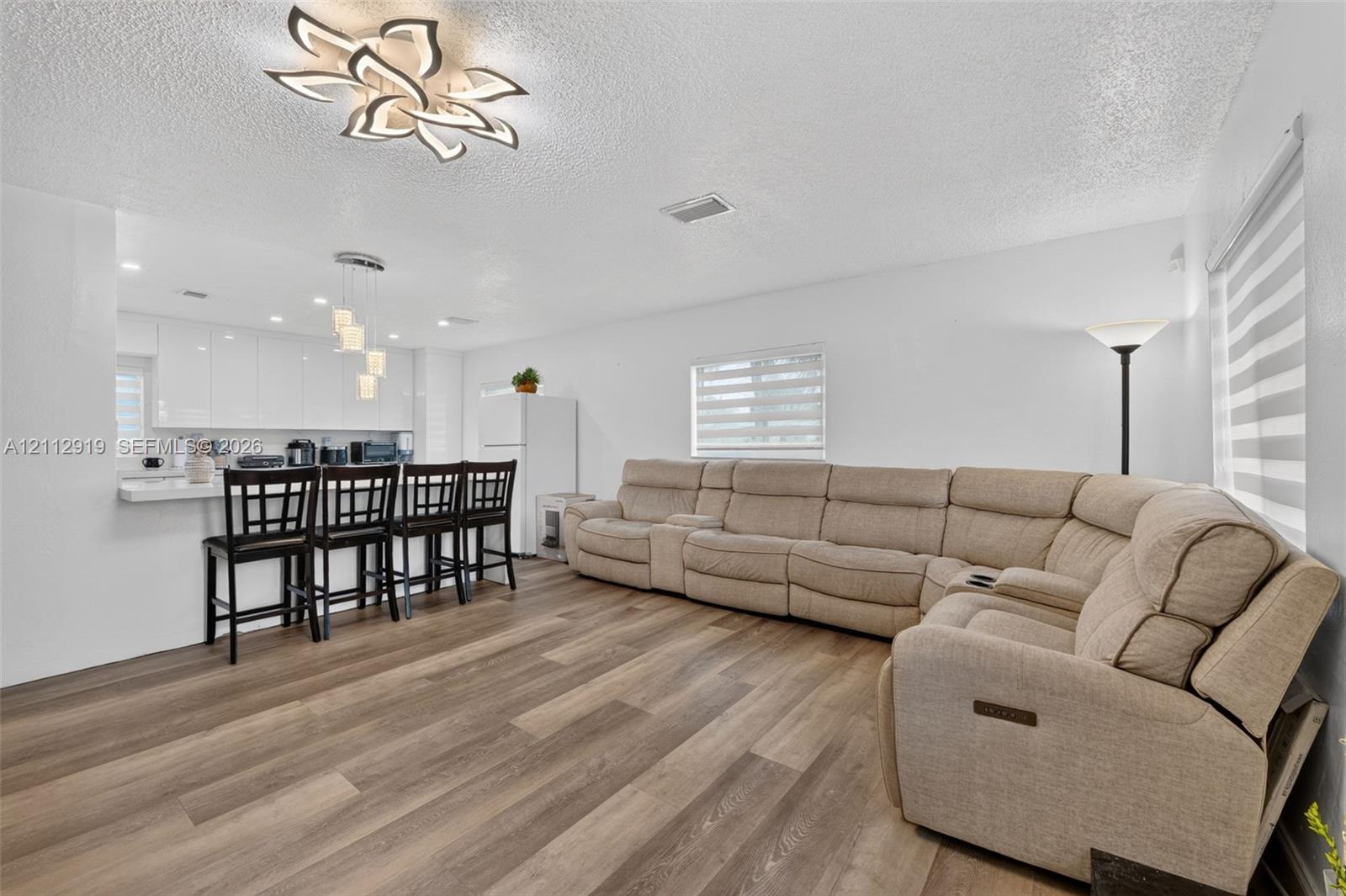 Open-concept interior featuring wood-finish flooring, a modern geometric ceiling light fixture, high-gloss white cabinetry, and multi-layered window shades