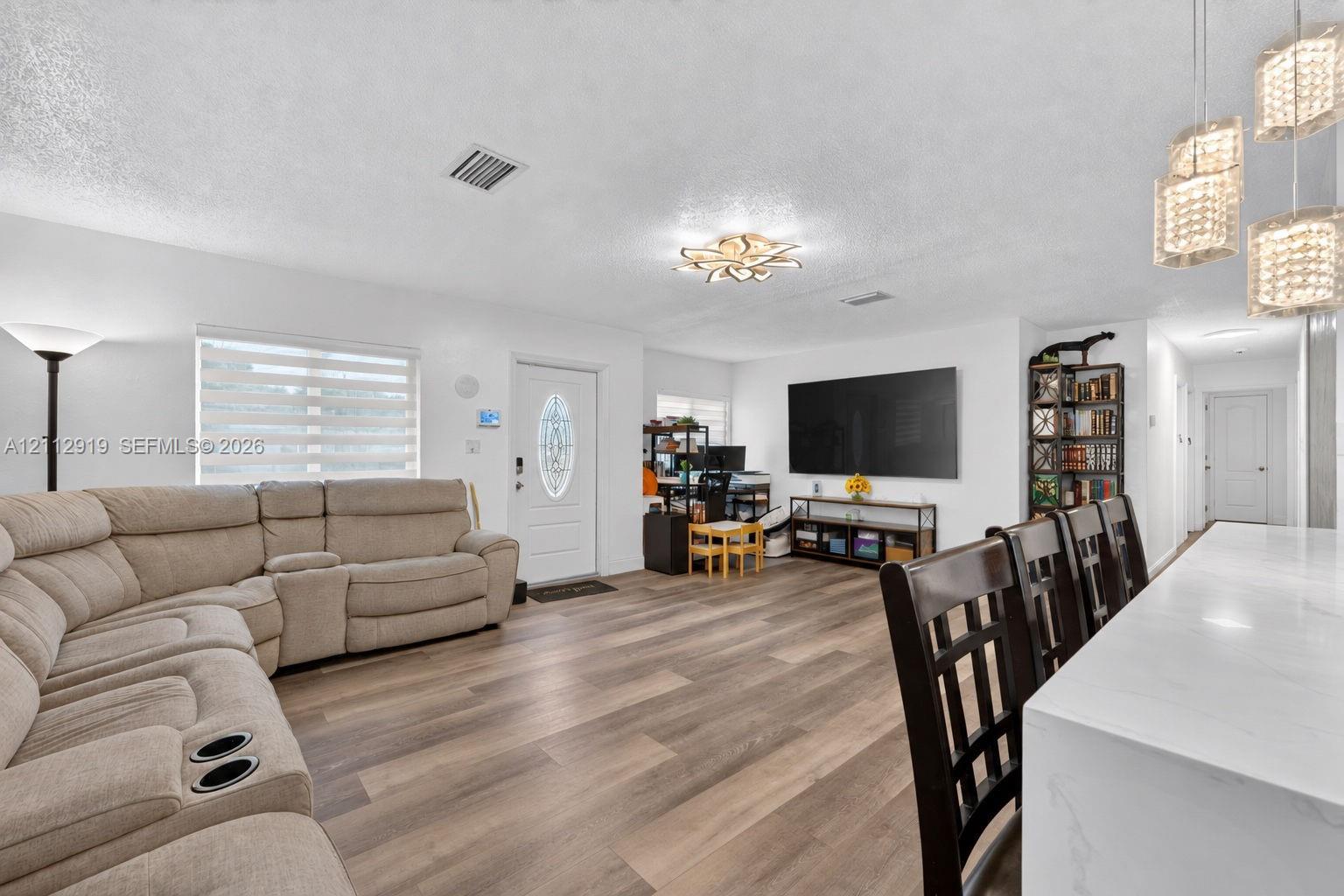 Open-concept interior featuring wood-finish flooring, multi-light pendant fixtures, a modern floral-inspired ceiling light, and an exterior door with decorative glass insert