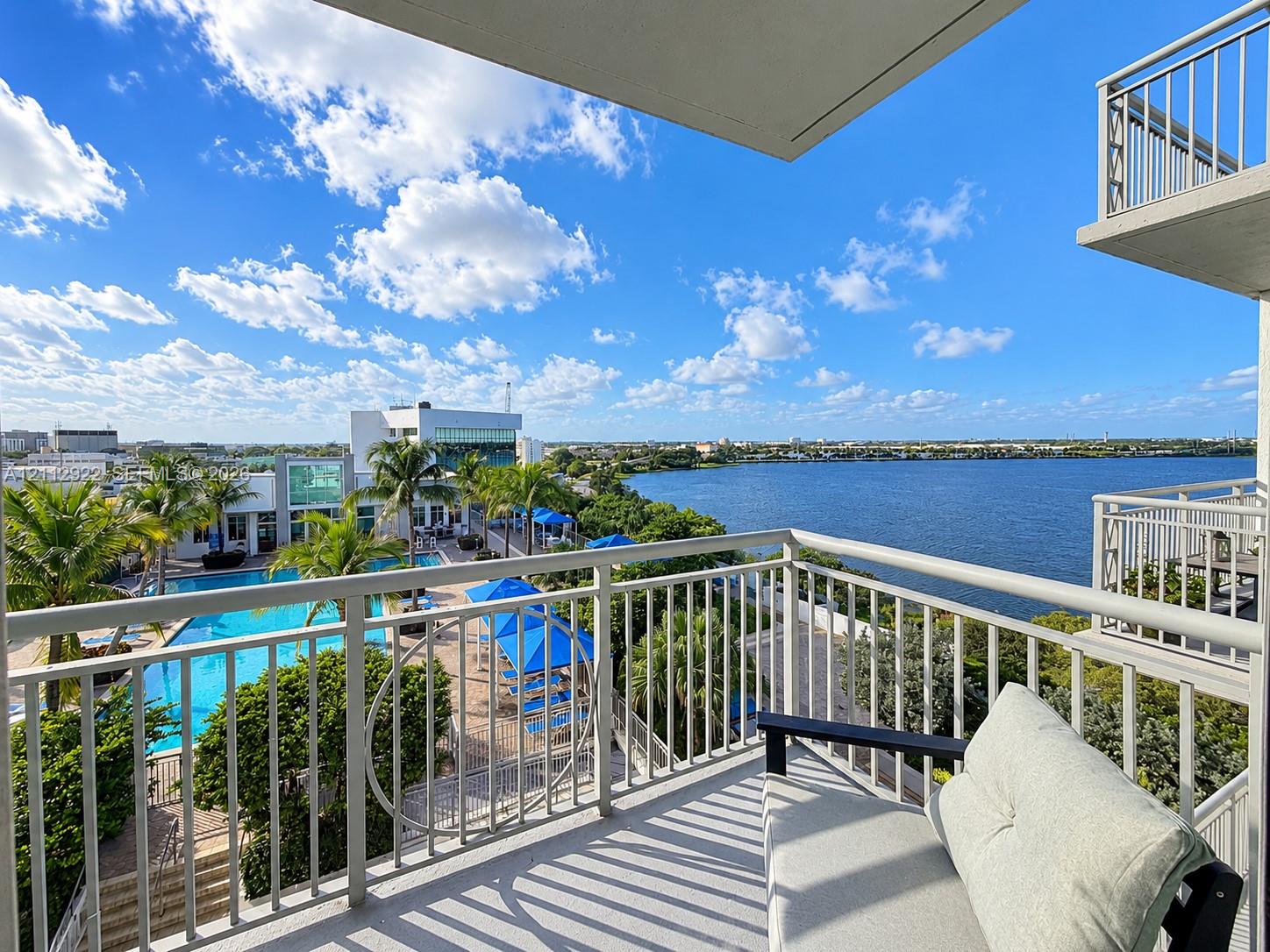 Private balcony featuring metal railings and concrete flooring, overlooking a swimming pool, mature palm trees, and waterfront scenery Private balcony featuring metal railings and concrete flooring, overlooking a swimming pool, mature palm trees, and waterfront scenery