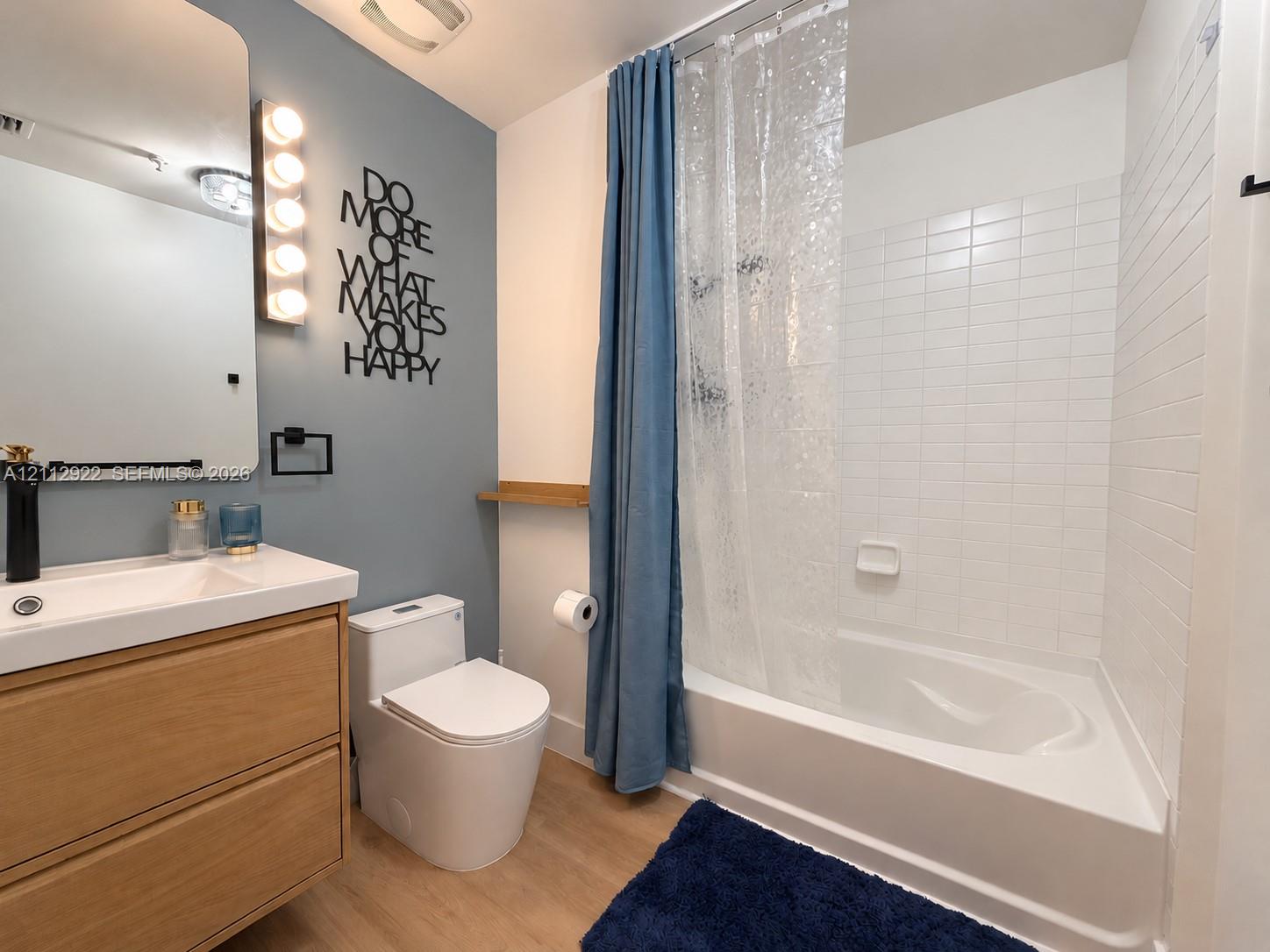 Bathroom featuring a wood-finish vanity with a rectangular sink, wall-mounted light fixture, mirror with rounded corners, and a tiled shower-tub combination