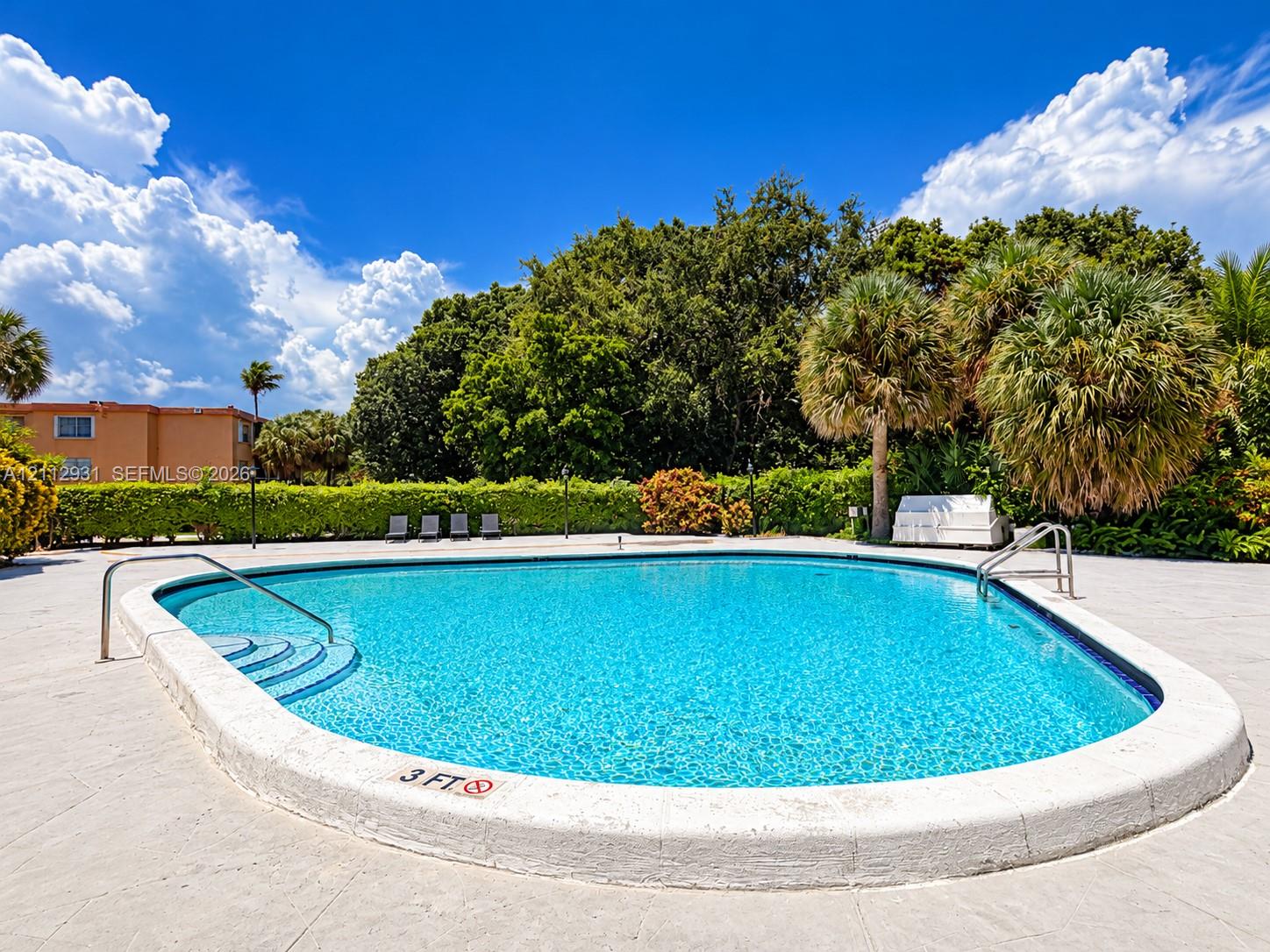 Outdoor swimming pool featuring a curved concrete deck, built-in steps with metal handrails, and surrounding mature landscaping with palm trees Outdoor swimming pool featuring a curved concrete deck, built-in steps with metal handrails, and surrounding mature landscaping with palm trees