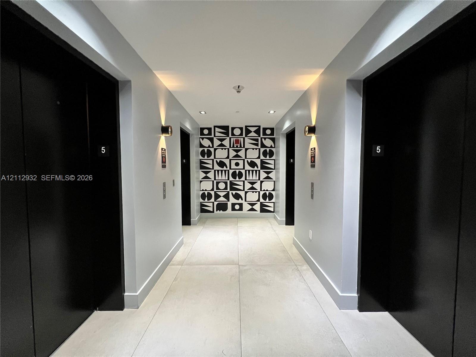 Hallway featuring large-format light-toned floor tiling, matte black door finishes, geometric decorative wall paneling, and wall-mounted cylindrical sconces
