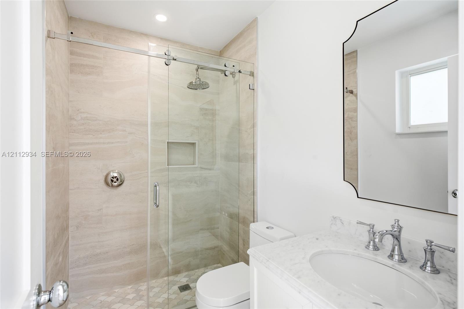 Walk-in shower with sliding glass door, stone-patterned wall tile, built-in recessed niche, rainfall showerhead, and stone vanity countertop with undermount sink