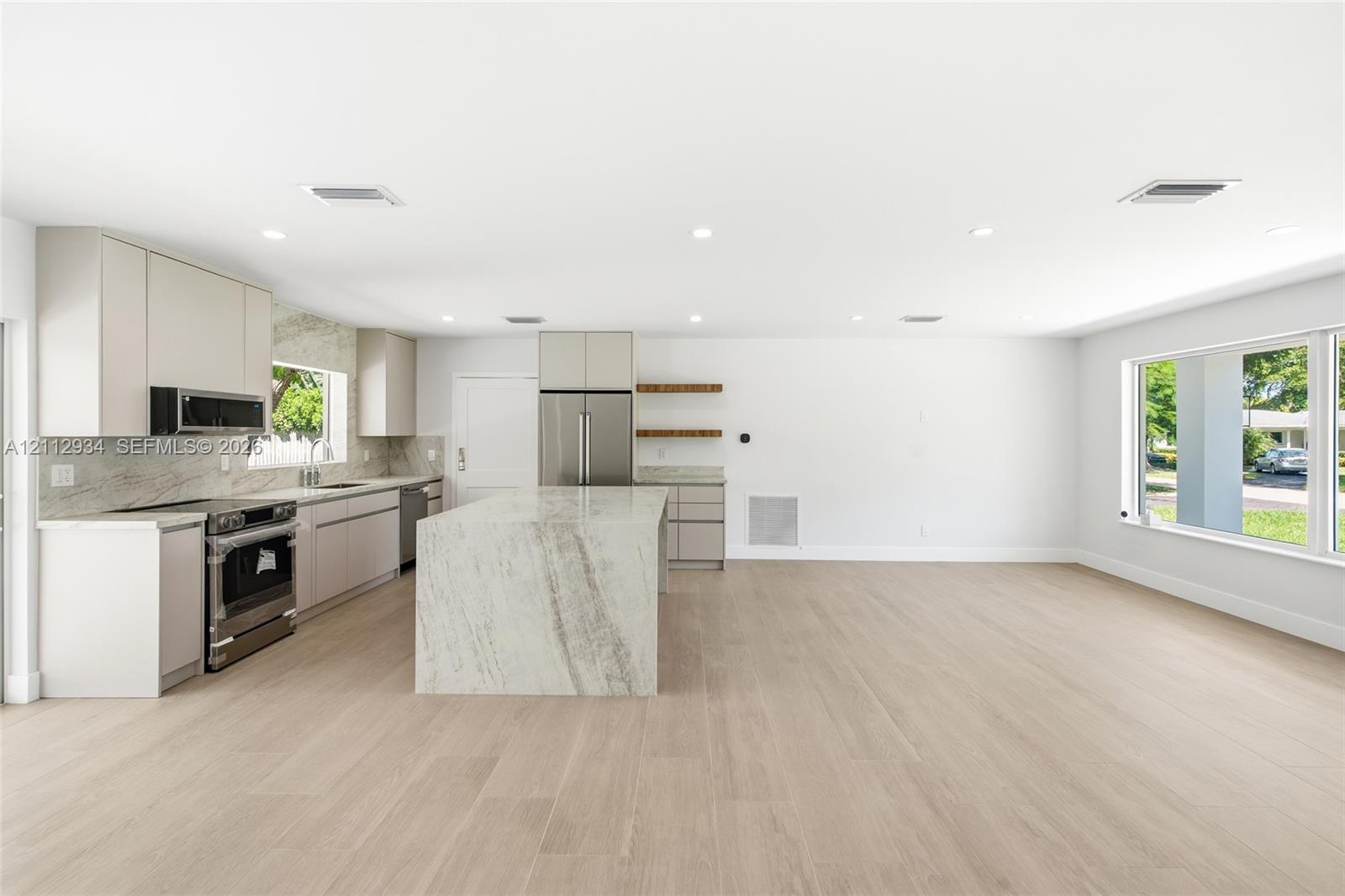 Kitchen featuring flat-panel cabinetry, a central island with stone-finish waterfall countertop, stainless steel appliances, wood-finish flooring, and floating shelves